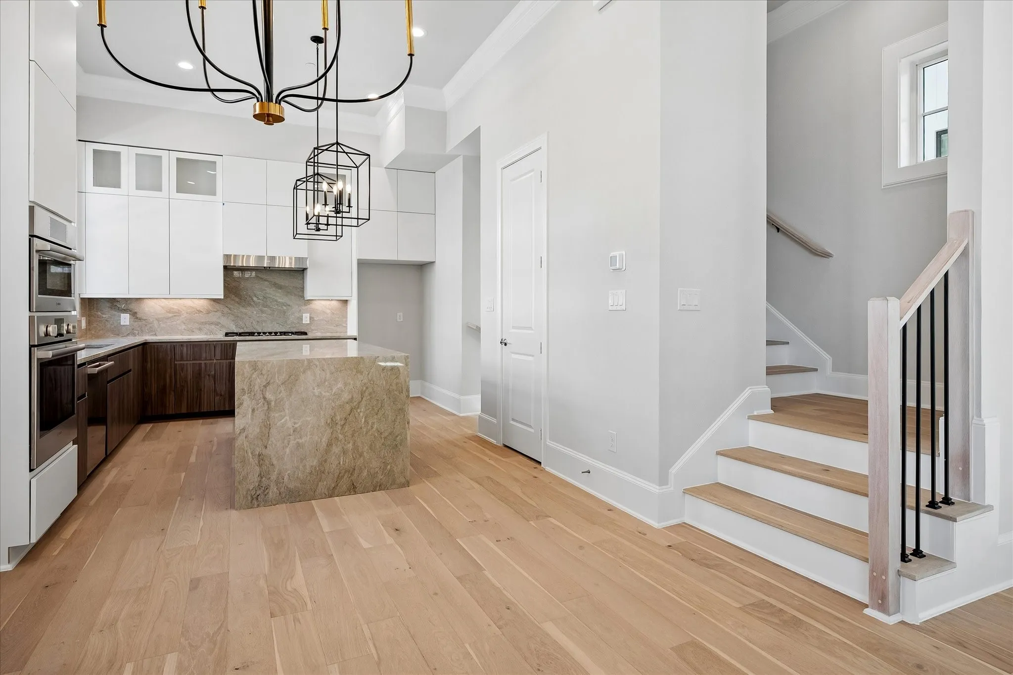 Open-concept kitchen and dining area featuring white cabinetry with stainless steel appliances, a waterfall-edge island with natural stone countertop, wide-plank wood-finish flooring, and a geometric pendant light fixture