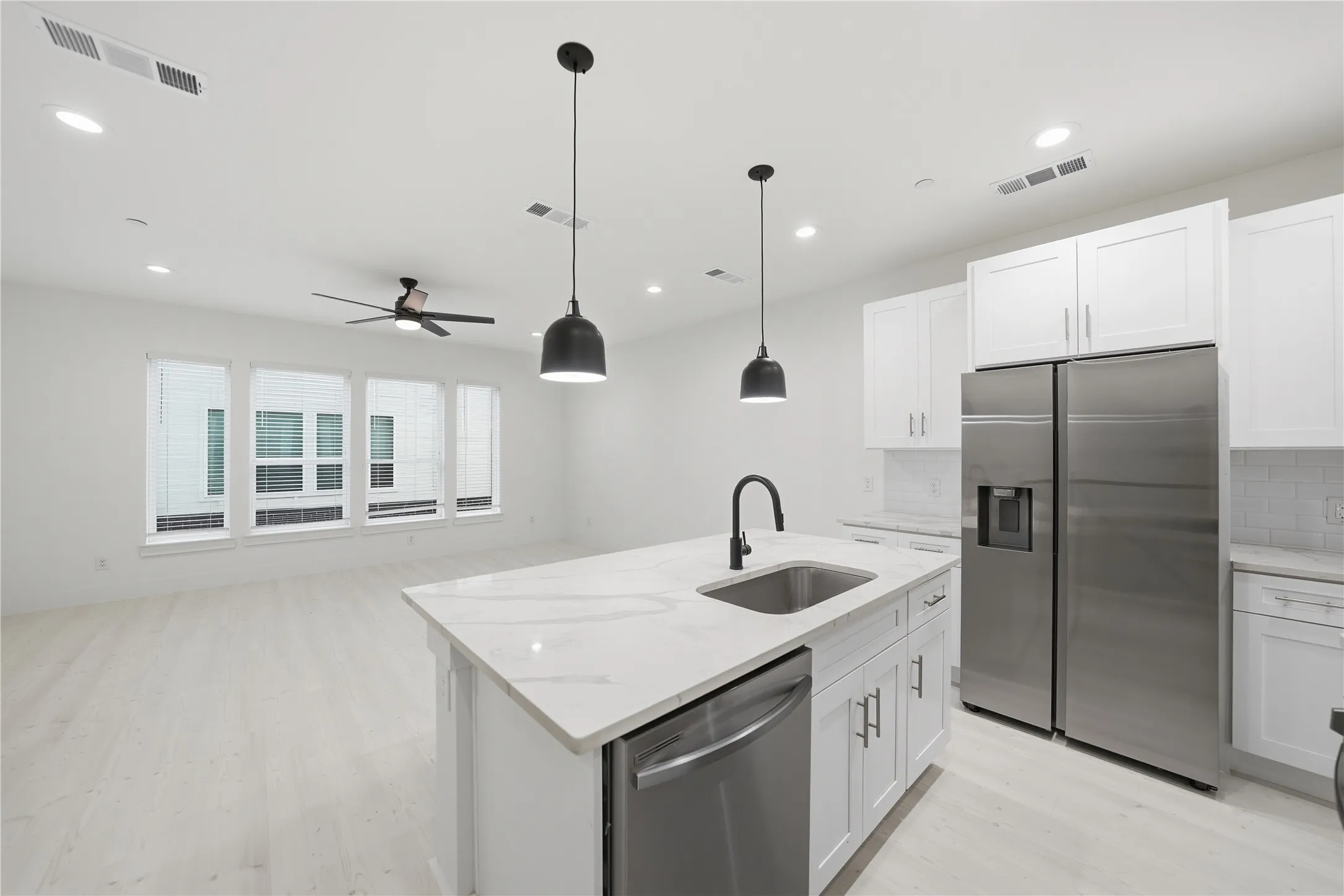 Open-concept kitchen and living area featuring a central island with integrated sink, white shaker cabinetry, stainless steel appliances, and light wood-finish flooring