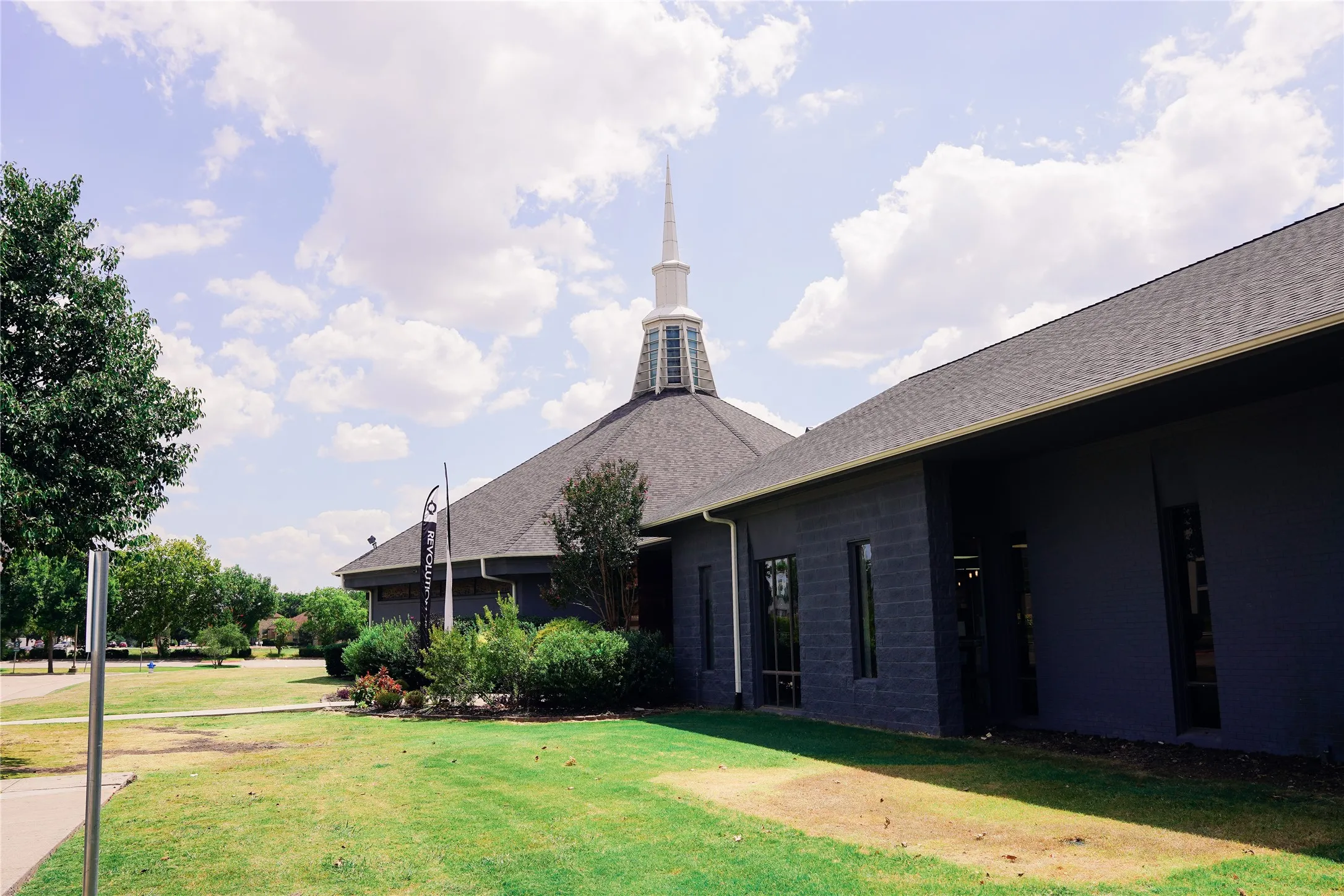 Pitched shingle roof with a prominent white steeple, exterior walls finished in dark gray, and surrounding green landscaping