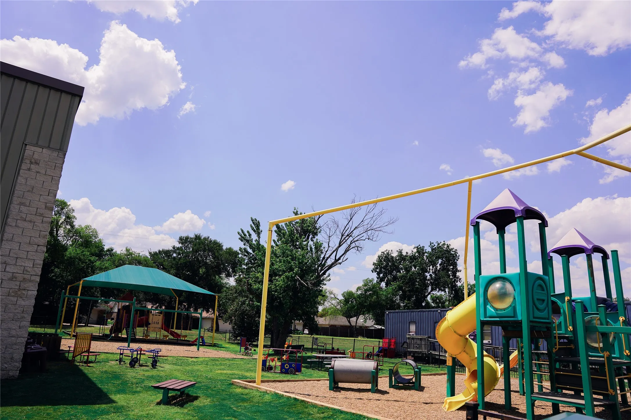 Outdoor recreational area featuring multiple playground structures, a shaded play space, a manicured lawn, and mature trees