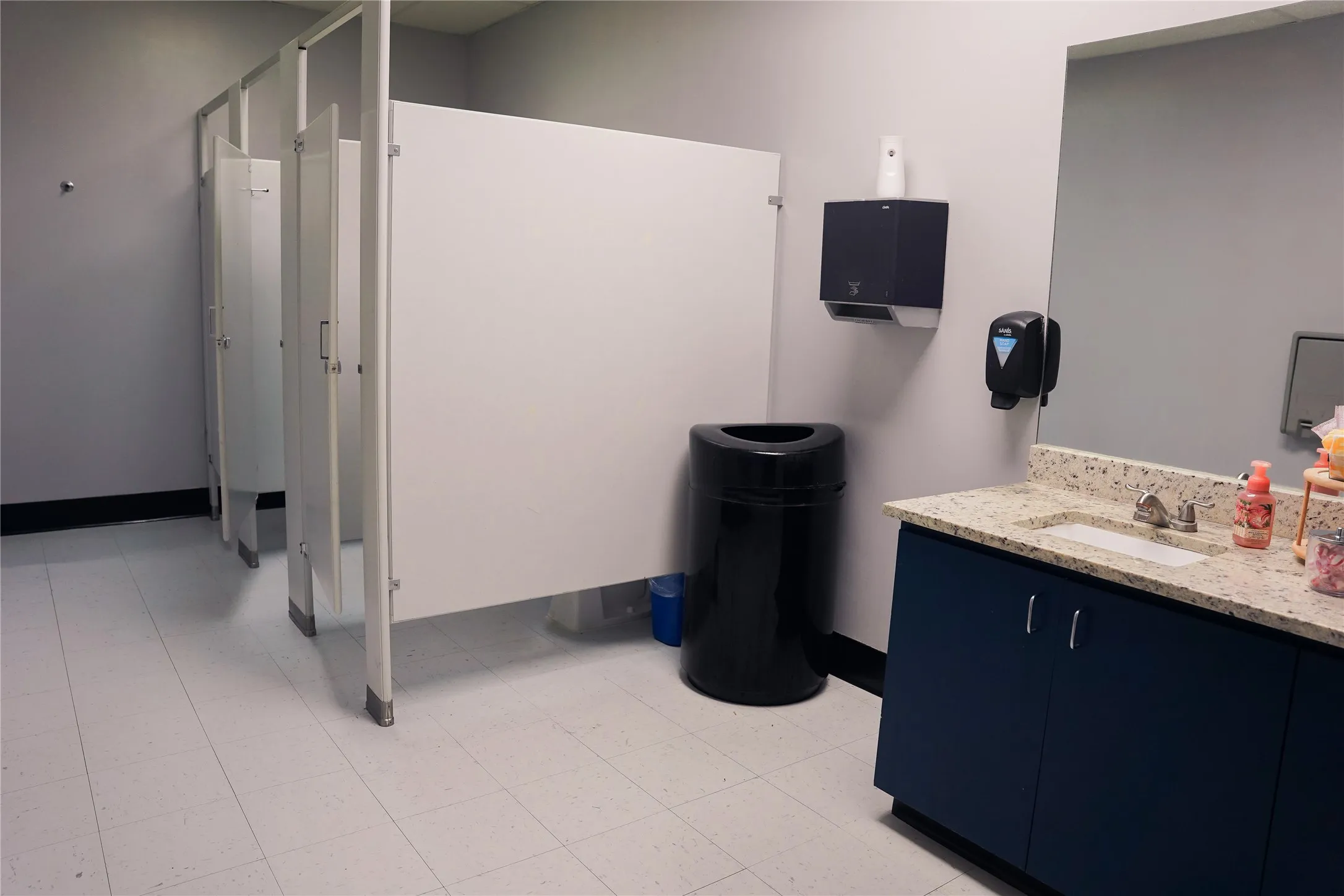 Restroom featuring multiple stalls with privacy panels, a vanity with a light-colored countertop, an integrated sink basin, and a wall-mounted mirror