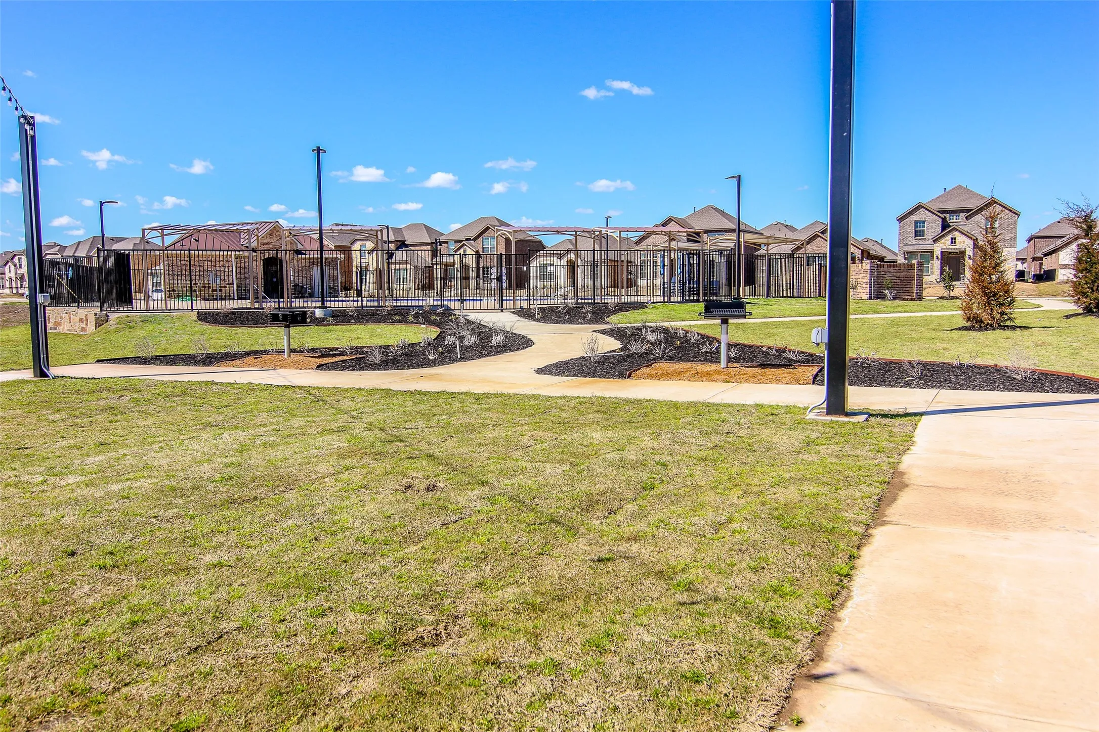 Community green space featuring a paved walkway, established landscaping, and a fenced recreation area with built-in grills