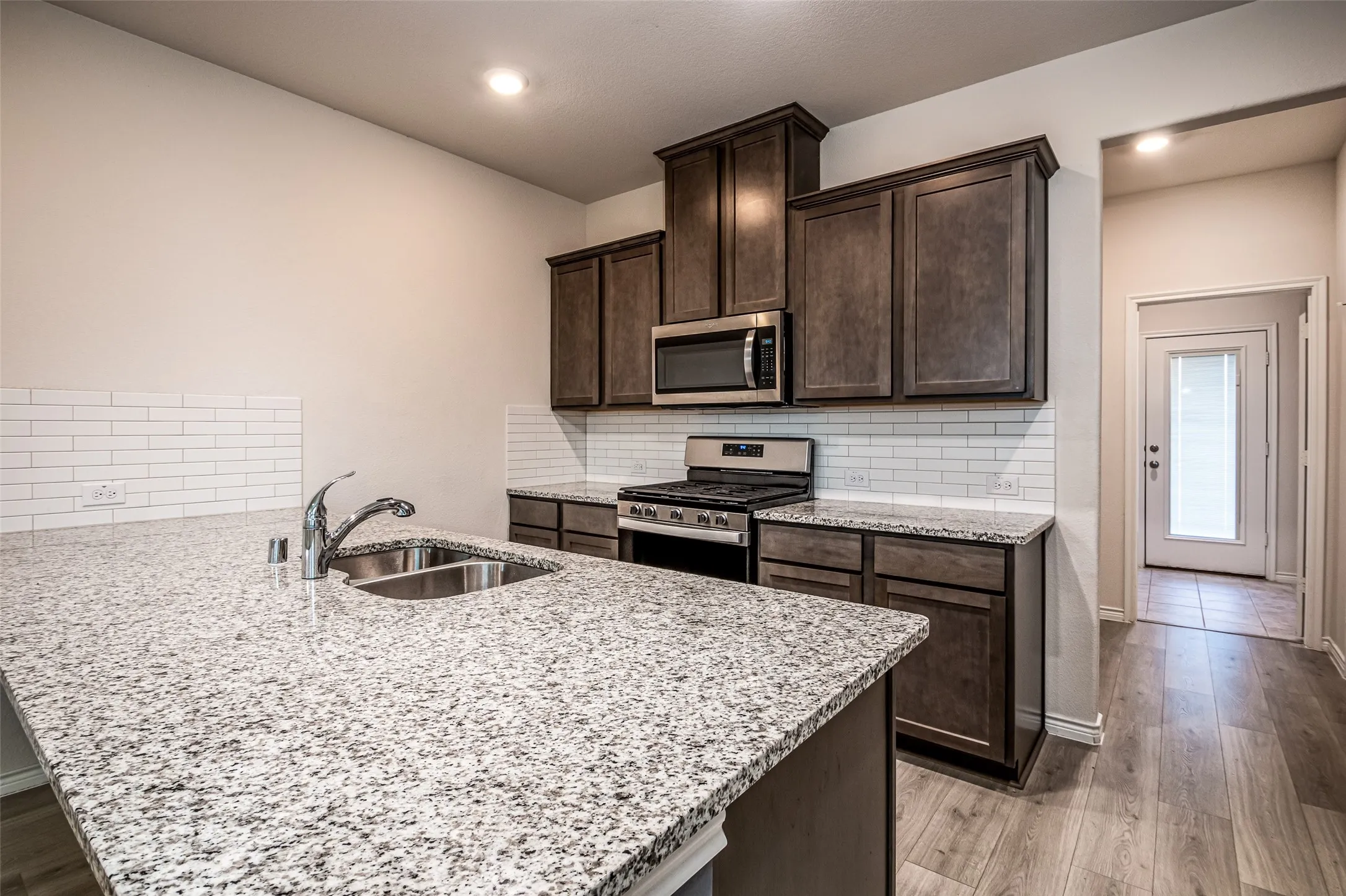 Kitchen featuring granite countertops, stainless steel appliances, dark wood cabinetry, a subway tile backsplash, and wood-finish flooring