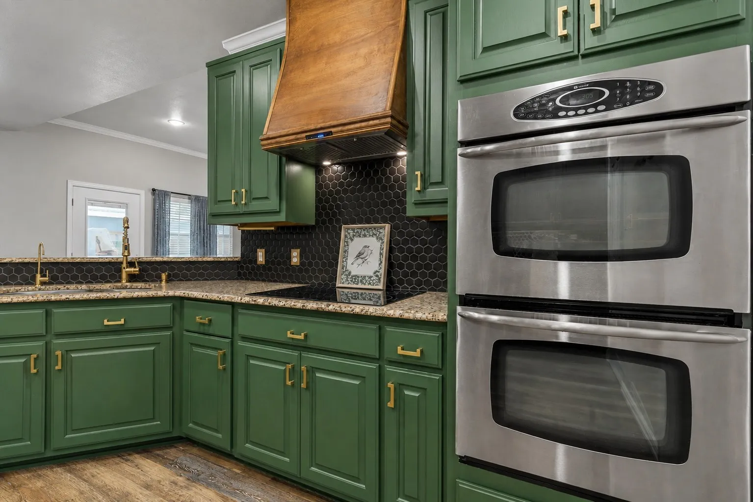 Green cabinetry with gold-tone hardware, wood-finish flooring, black hexagonal tile backsplash, light-toned speckled countertops, and stainless steel double ovens