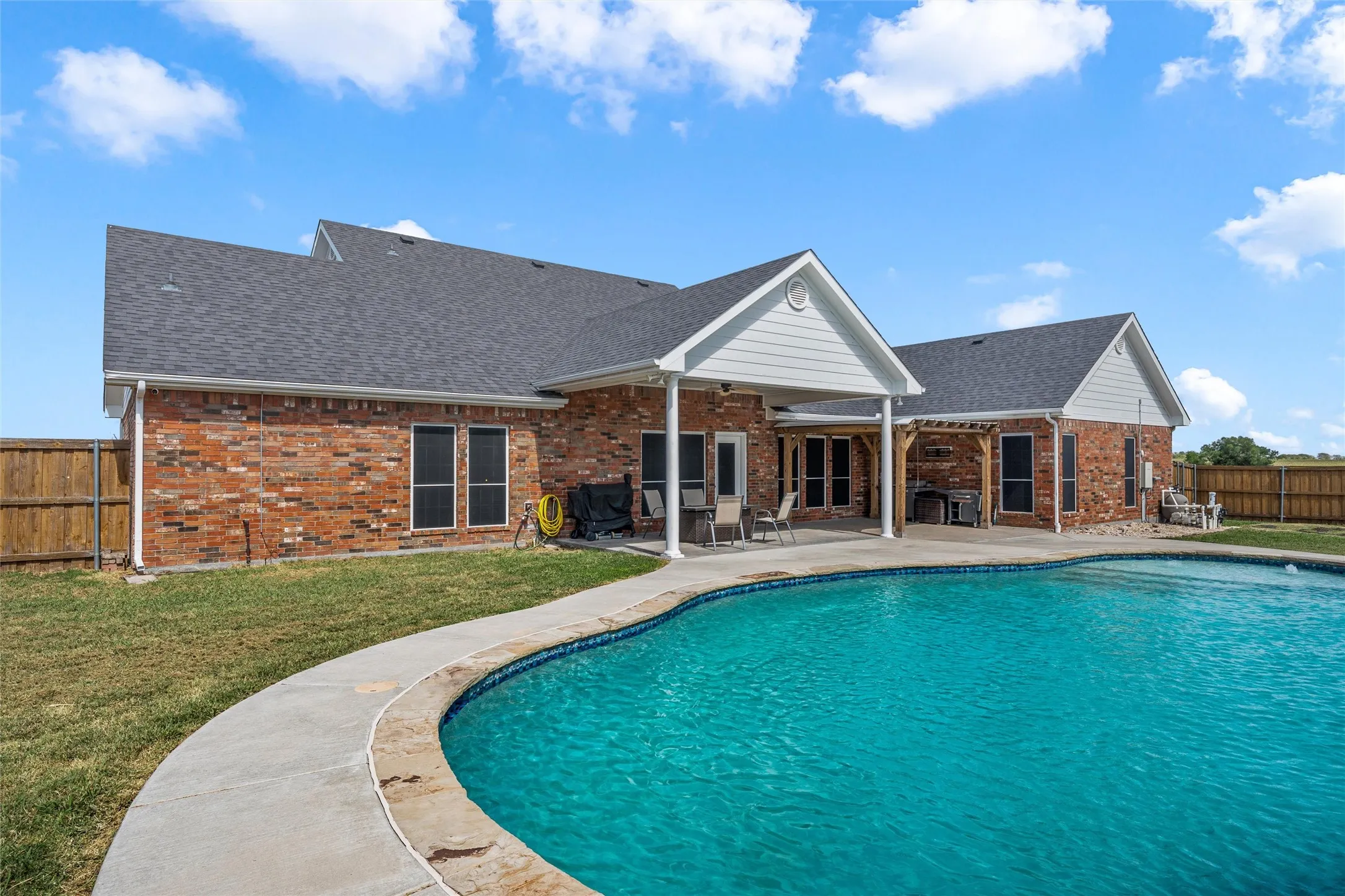 Brick exterior with a dark shingle roof, featuring an in-ground swimming pool with stone coping, a covered patio, and a wood privacy fence