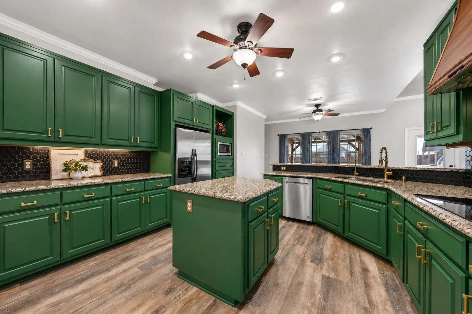 Chef's kitchen showcasing rich green cabinetry with brass hardware, granite countertops, a large center island with seating, stainless steel appliances, hexagonal tile backsplash, recessed and ceiling fan lighting, and wood-finish flooring throughout the open layout