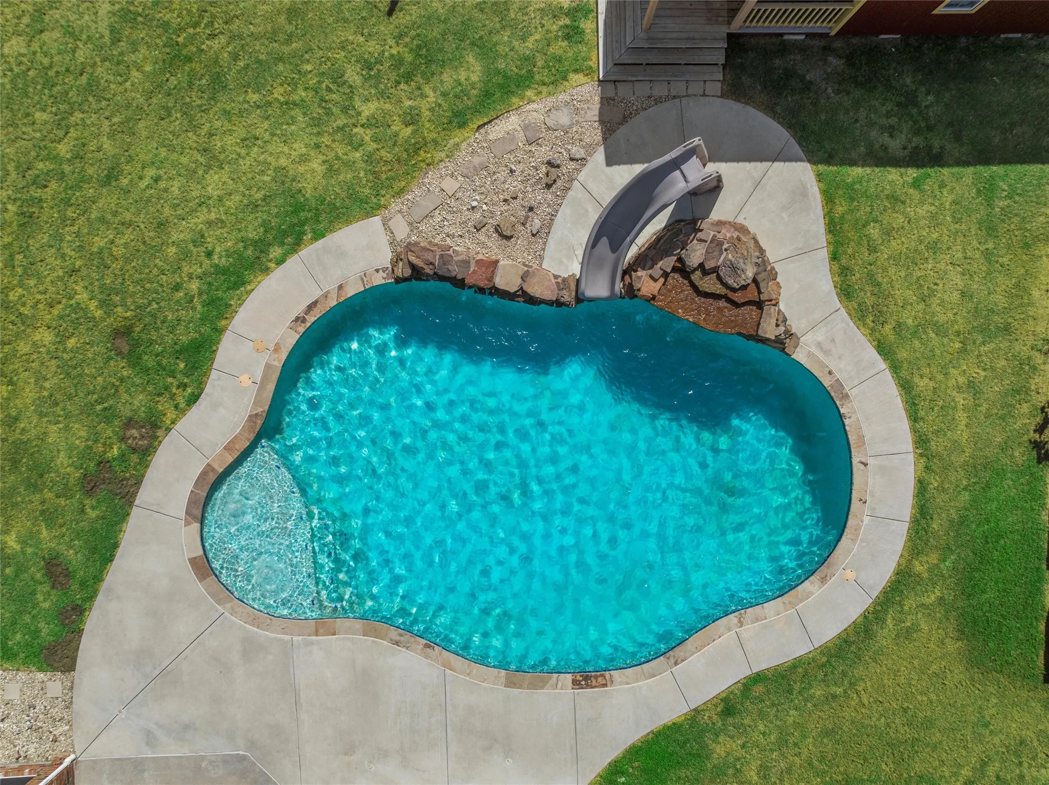 Freeform swimming pool with integrated slide, rock features, concrete decking, and surrounding green lawn