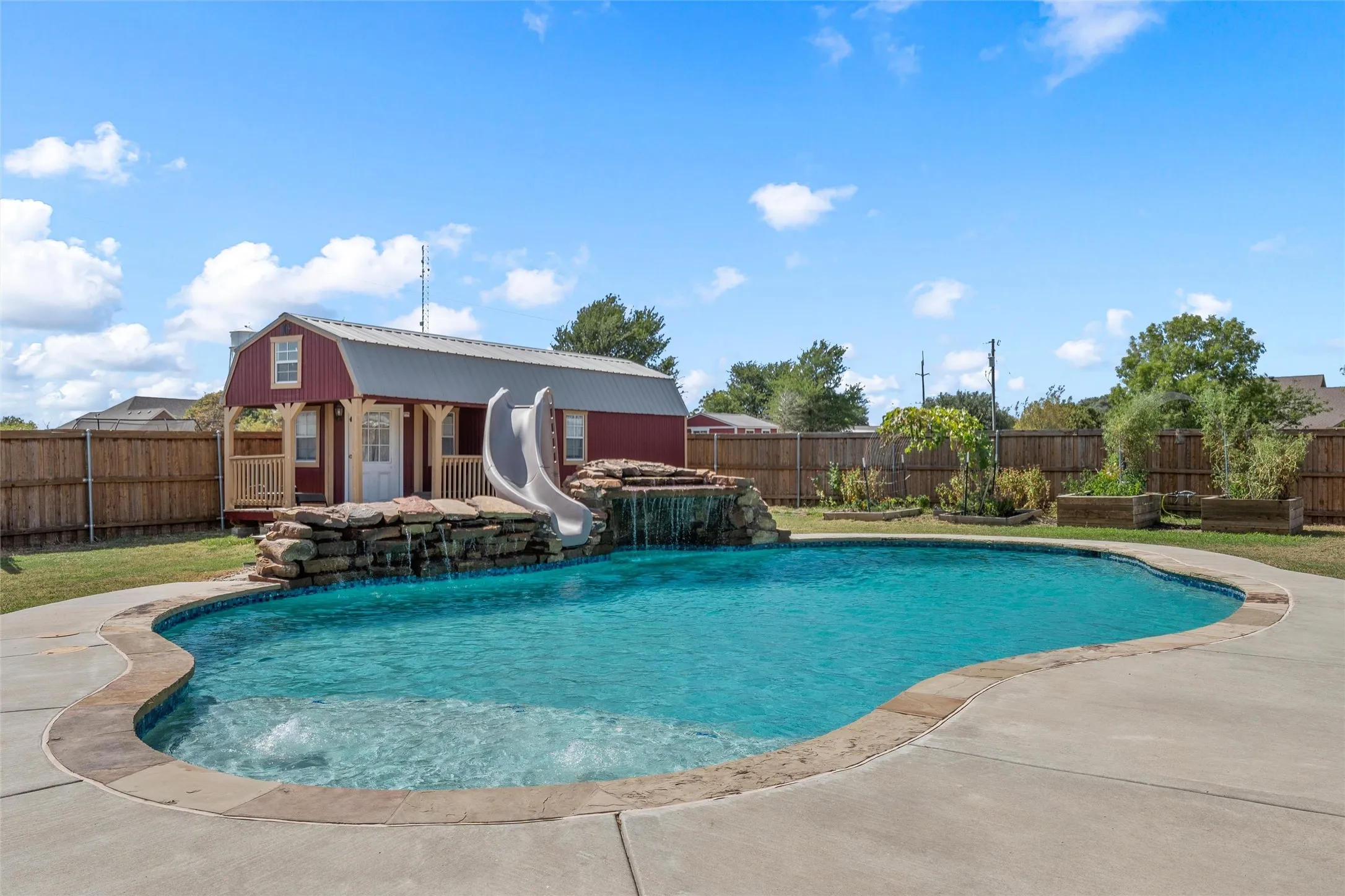Freeform swimming pool with rock waterfall feature, concrete decking, and fenced yard