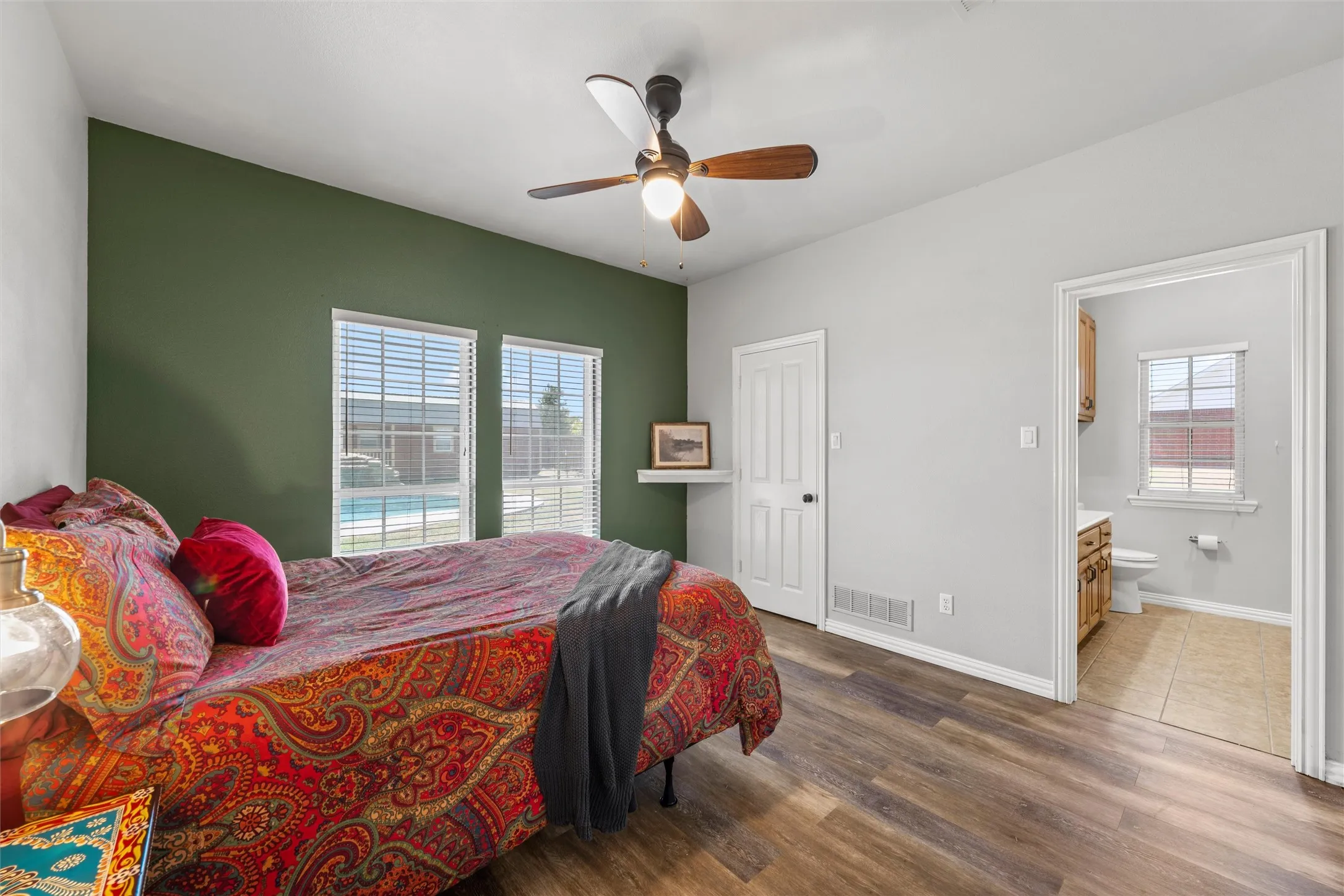 Wood-finish flooring, ceiling fan, two windows with blinds, recessed shelving, and an adjacent bathroom with tile flooring