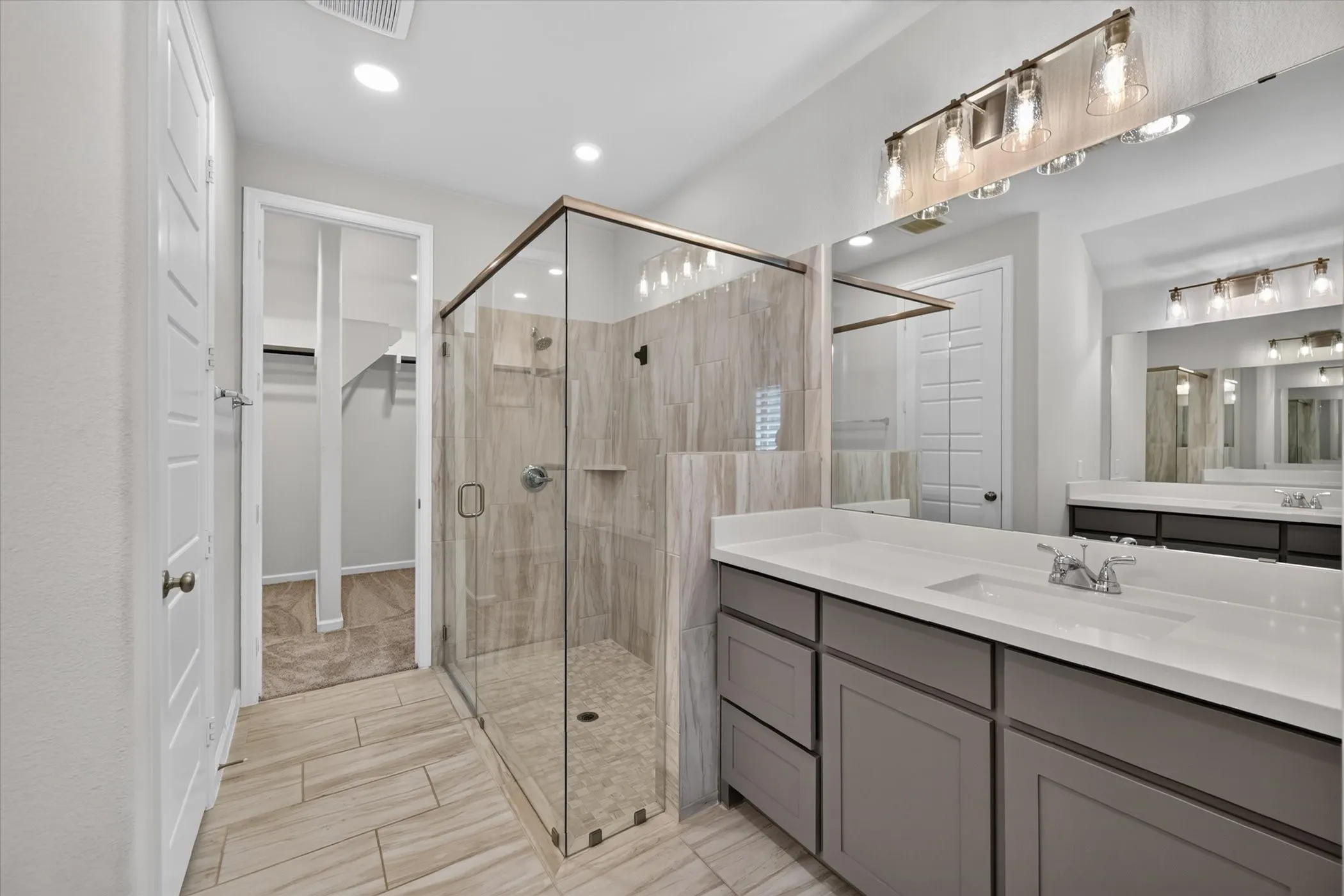Glass-enclosed shower with tile surround and floor, dual vanity with undermount sinks and white countertops, tile flooring, and overhead vanity lighting
