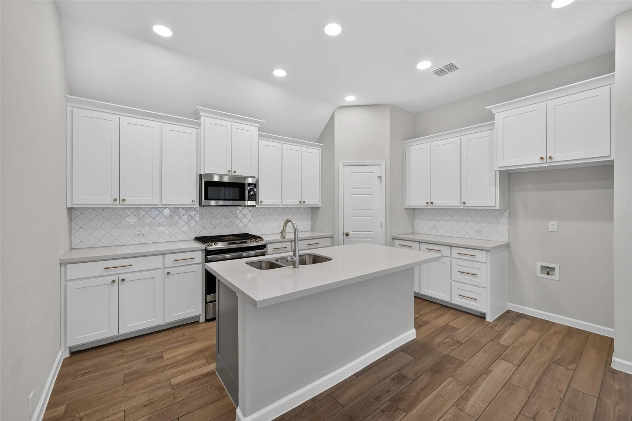 Kitchen featuring white cabinetry, a central island with an undermount sink, stainless steel range and microwave, tiled backsplash, and wood-finish flooring