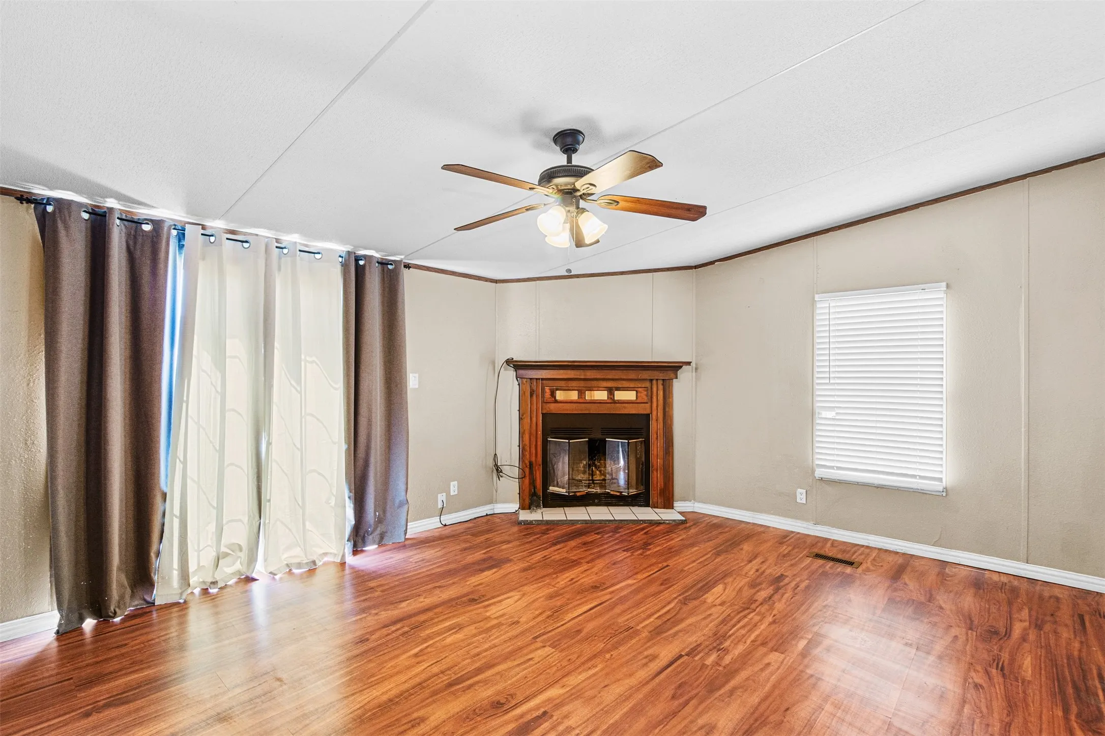 Wood-finish flooring, corner fireplace with wood mantel, ceiling fan, and window with blinds