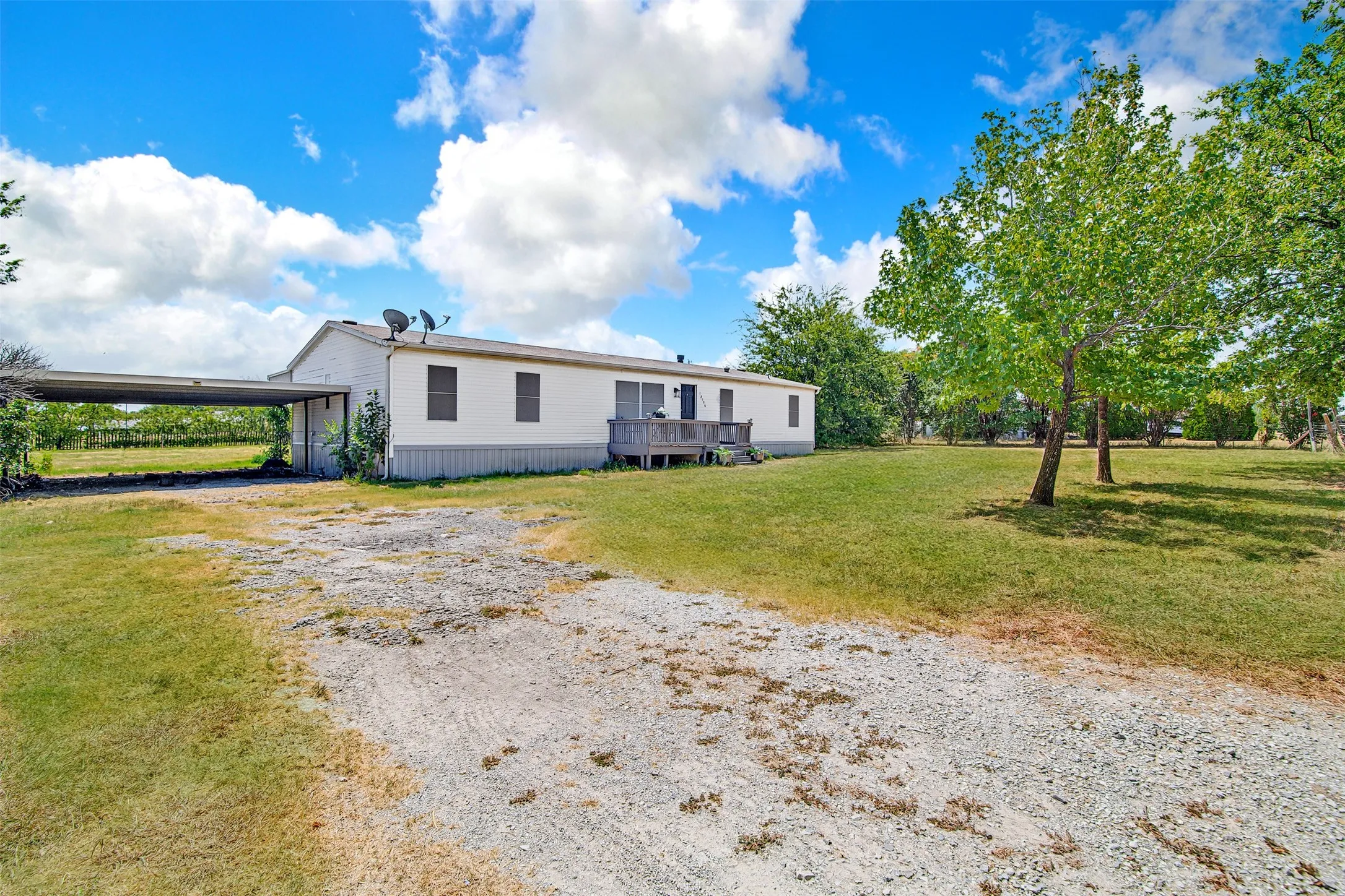 White exterior siding with dark window shutters, expansive green lawn with mature trees, and a gravel driveway