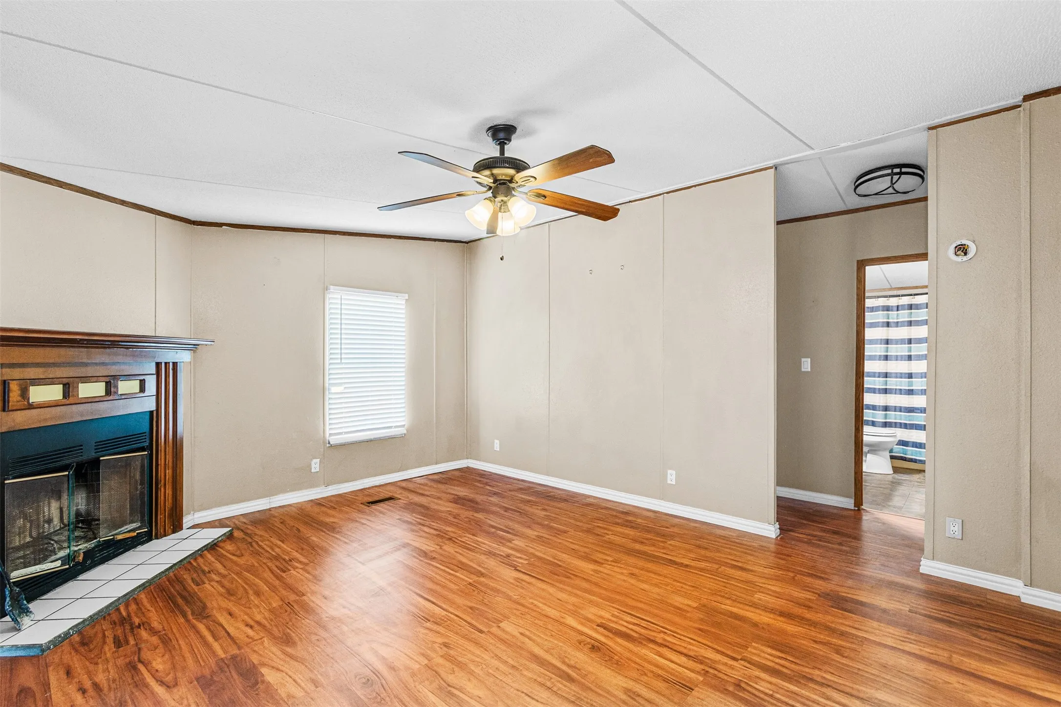 Wood-finish flooring throughout, ceiling fan with integrated lighting, built-in fireplace with a tiled hearth and wood-finished mantel, window with horizontal blinds, and a flush-mount ceiling light fixture