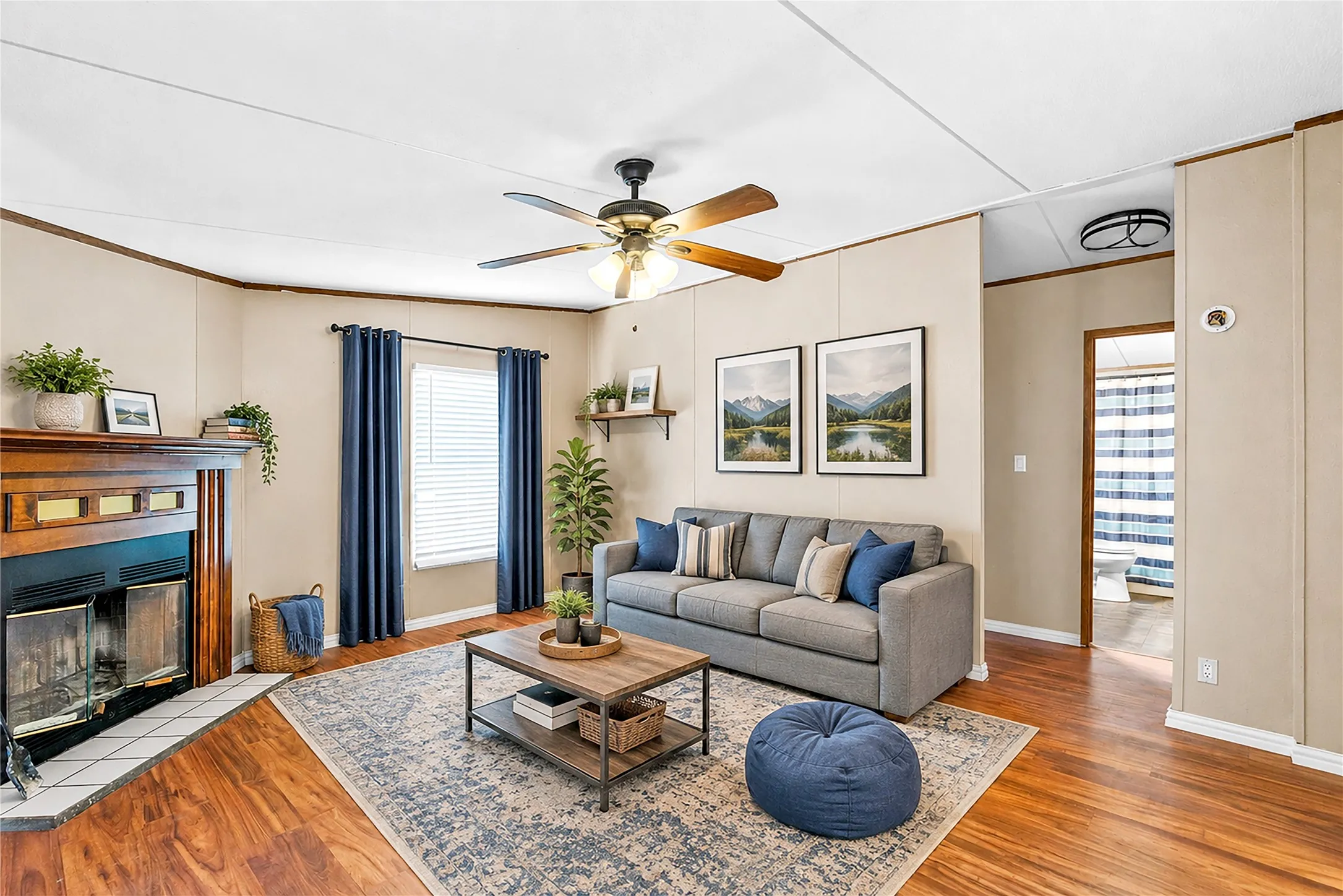 Living area featuring wood-finish flooring, a built-in fireplace with a tiled hearth, a ceiling fan with integrated lighting, and a window with blinds. Virtual staging photo.