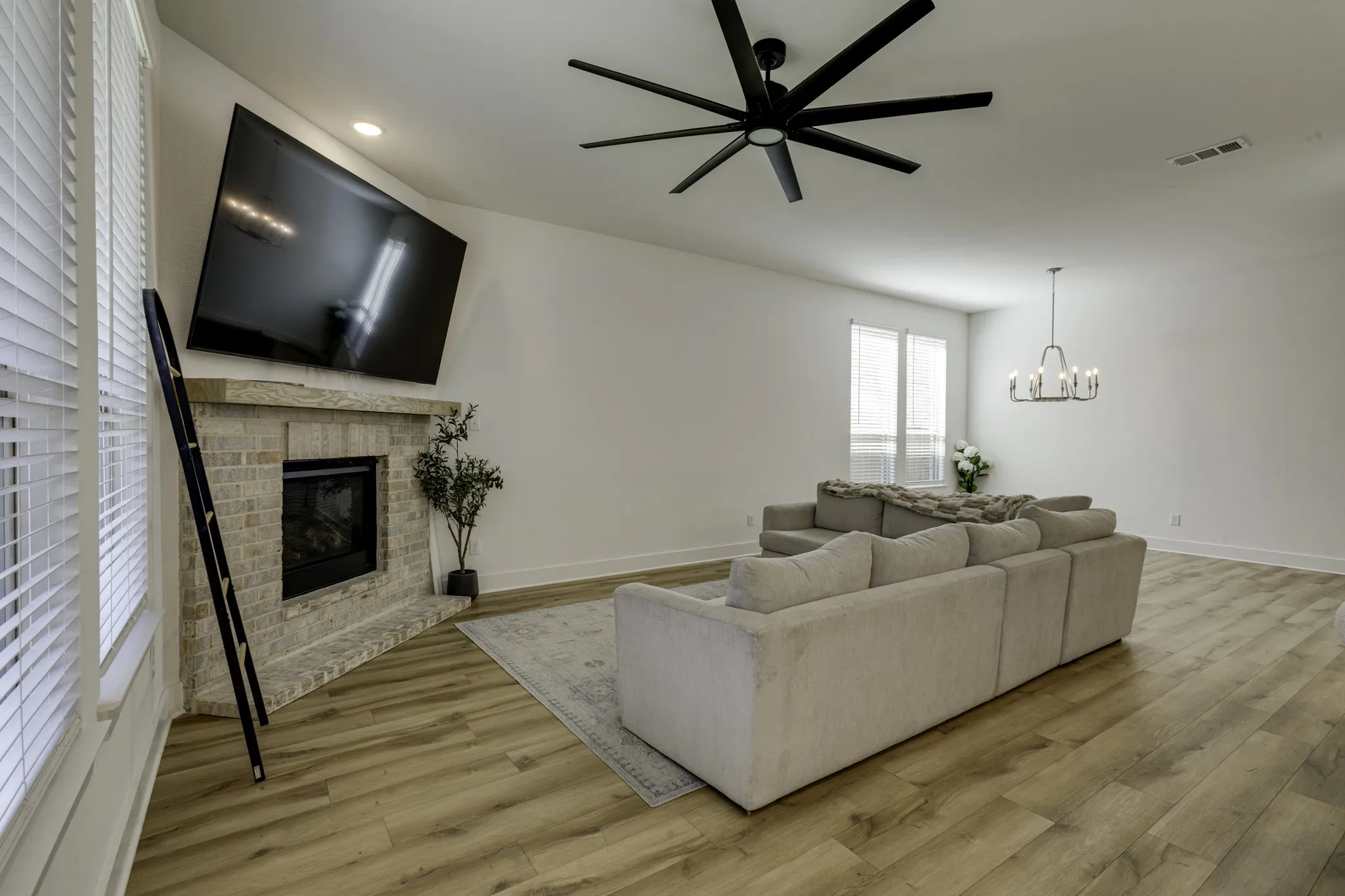 Living area featuring a corner fireplace with brick surround, wood-finish flooring, a large ceiling fan, a contemporary chandelier, and window blinds