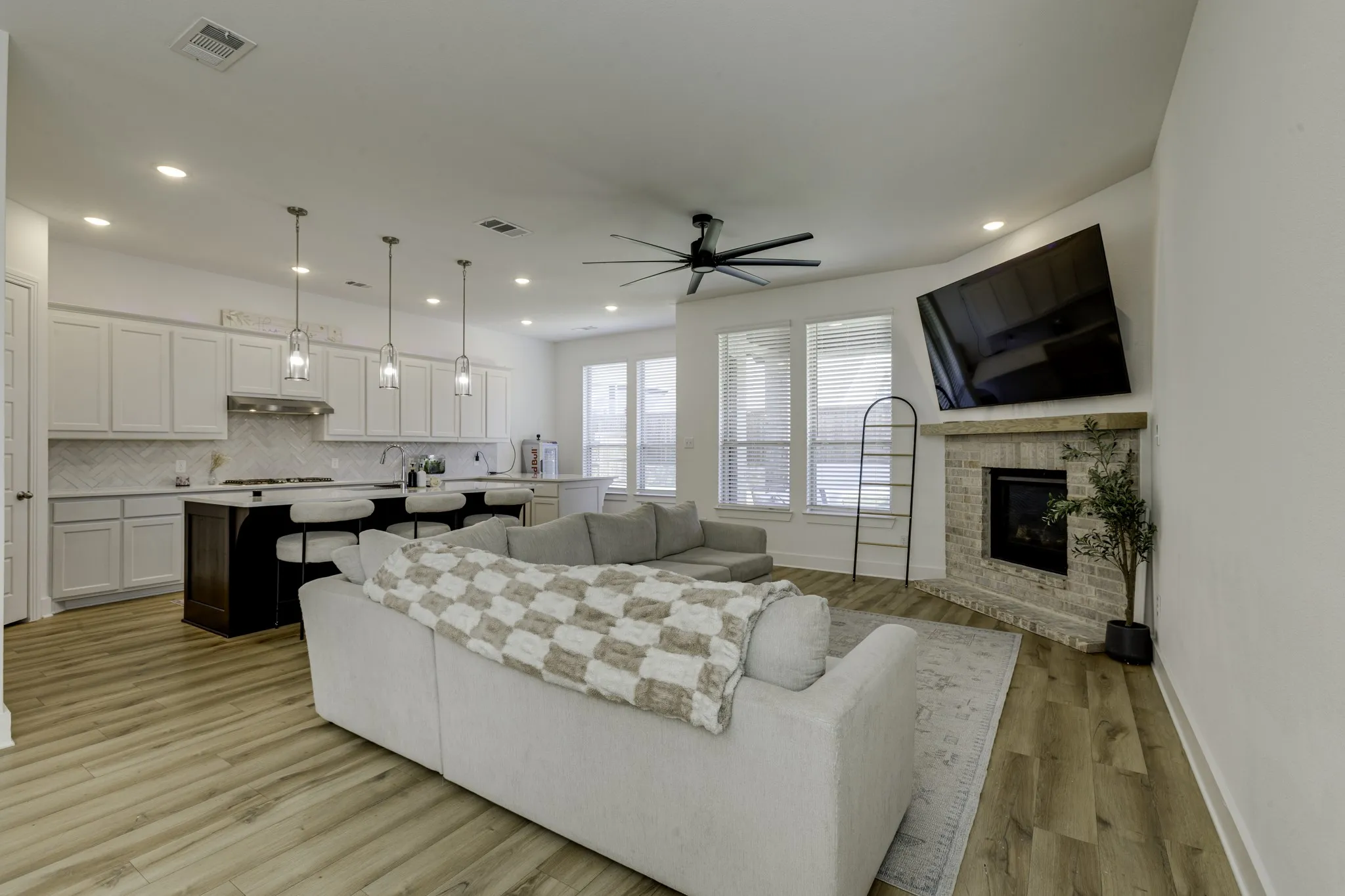 Open-concept living area featuring wood-finish flooring, a kitchen island with seating, pendant lighting, a ceiling fan, and a corner fireplace with a stone surround