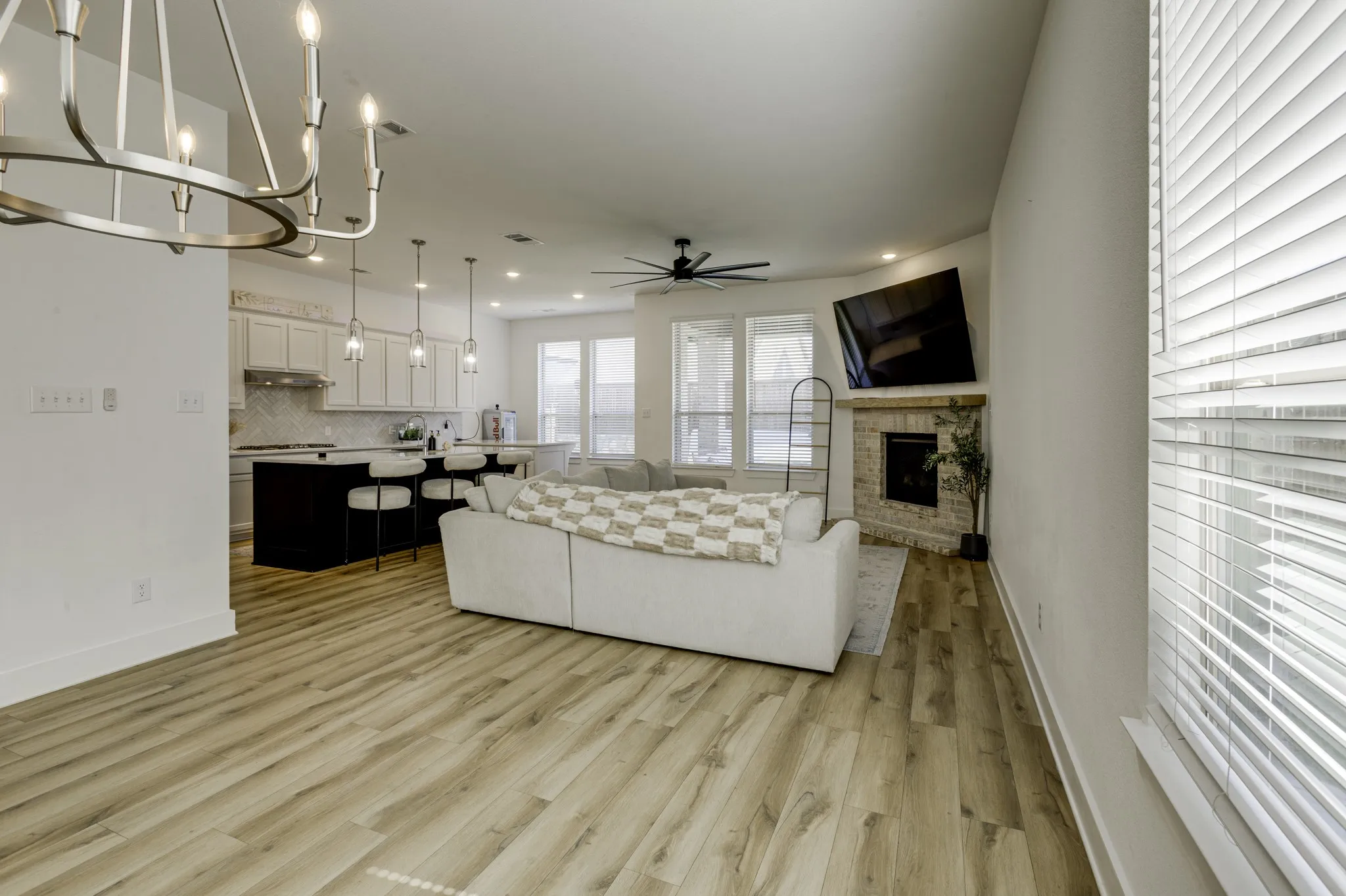 Open-concept living area featuring wood-finish flooring, a contemporary chandelier, a kitchen island with pendant lighting, a corner fireplace with a stone surround, and multiple windows