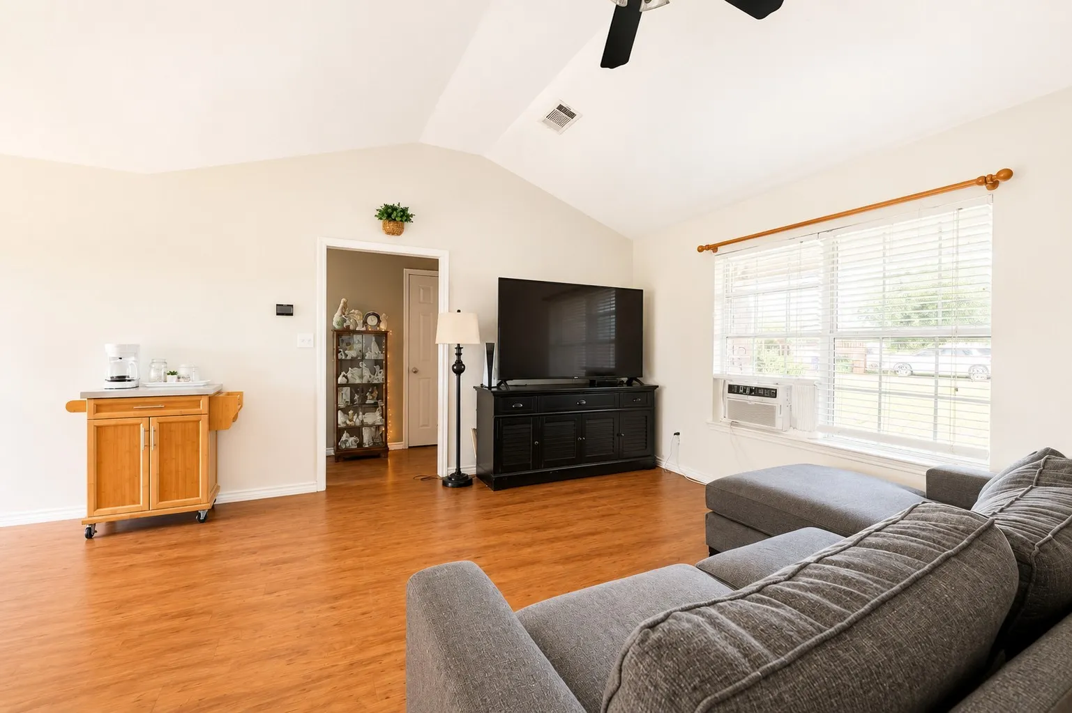 Vaulted ceiling, wood-finish flooring, white walls, large window with blinds, ceiling fan