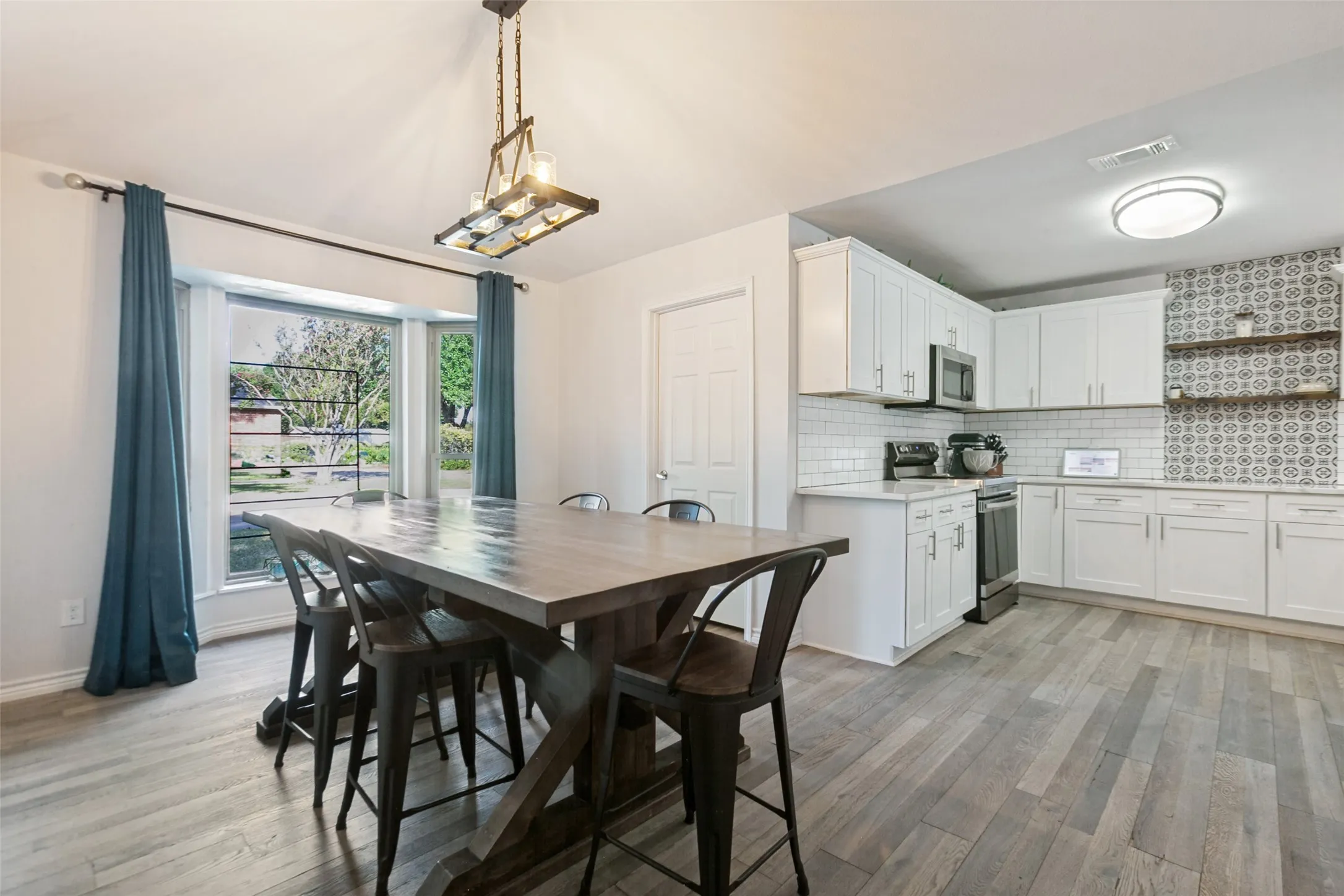 Open-concept kitchen and dining area featuring white cabinetry with stainless steel appliances, patterned tile backsplash, wood-finish flooring throughout, and large windows with blue drapery overlooking landscaped grounds