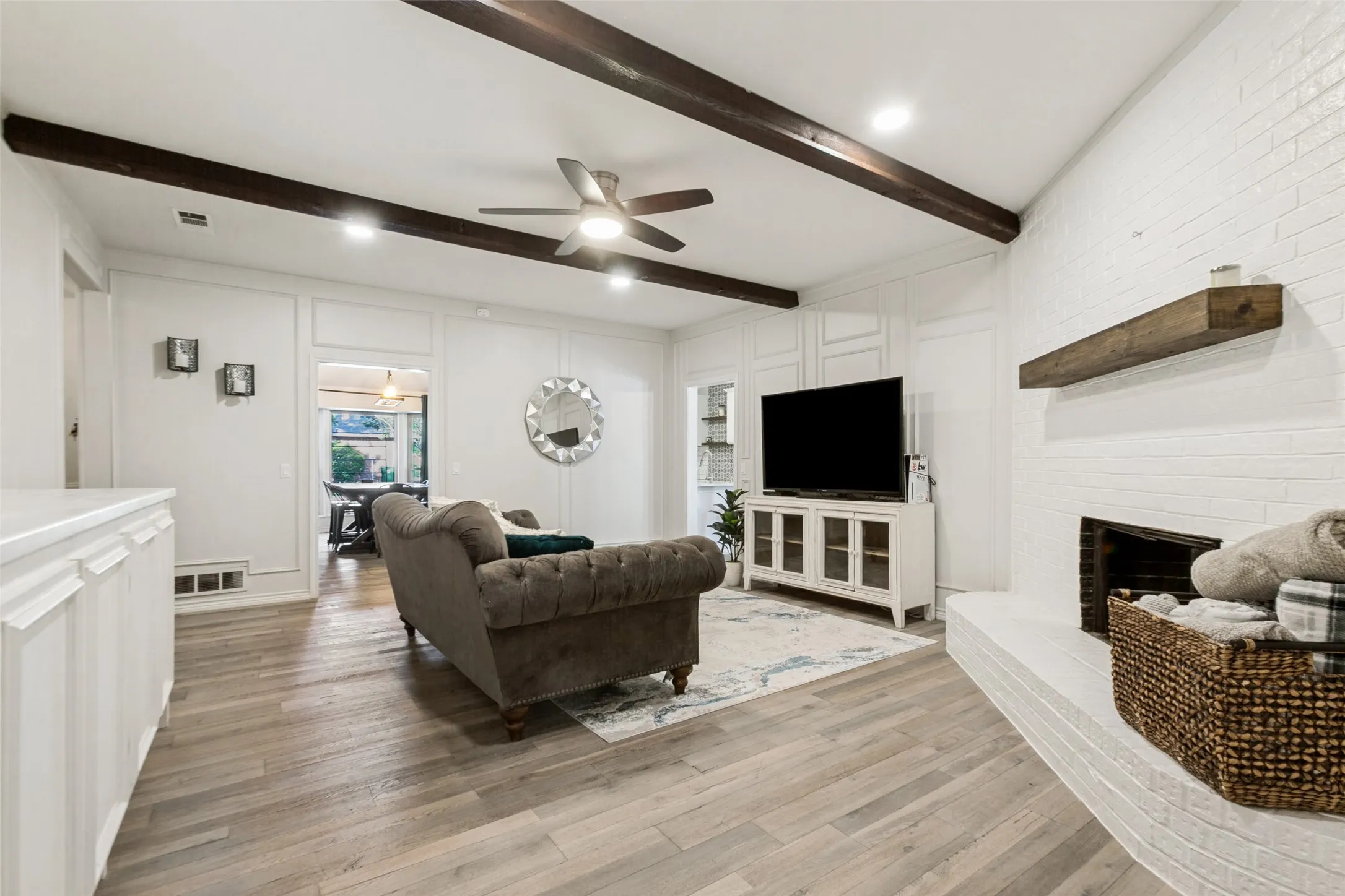 Wood-finish flooring, white brick fireplace with a wood mantle, white paneled wall accents, recessed lighting, and exposed ceiling beams