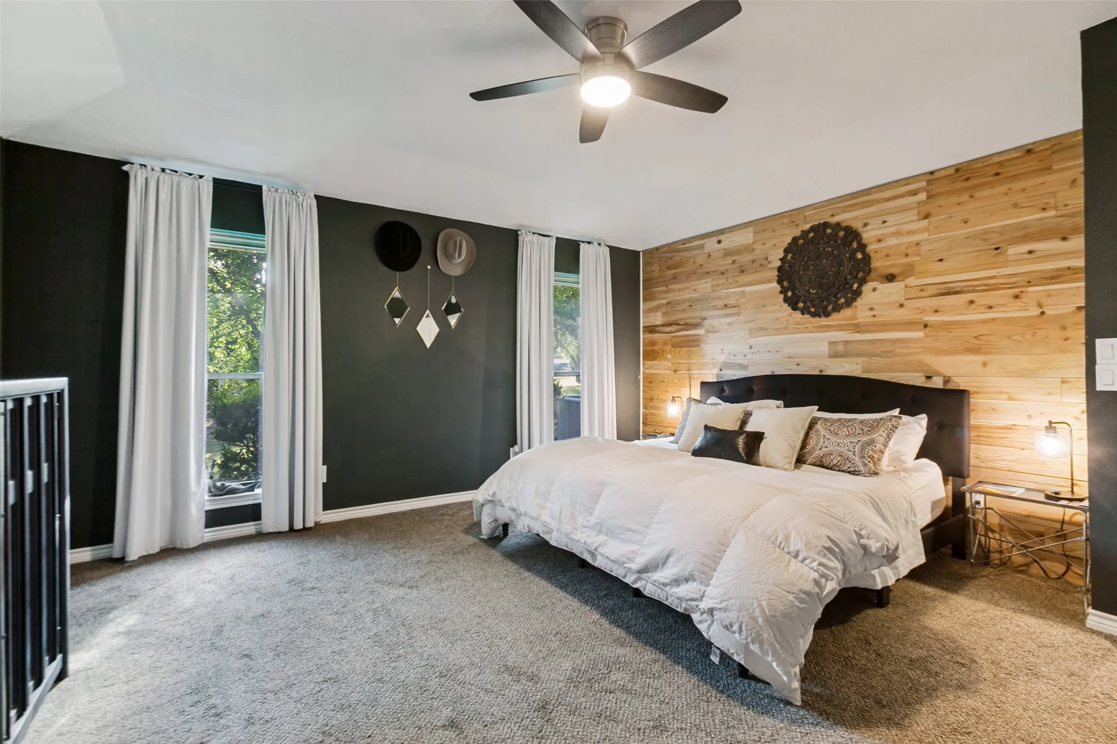 Primary Bedroom Suite with Accent Wood-Finish Wall Paneling, Ceiling Fan with Integrated Lighting, Black Metal Bed Frame, Dual Glass Doors Opening to Natural Light, and Plush Carpeting Throughout