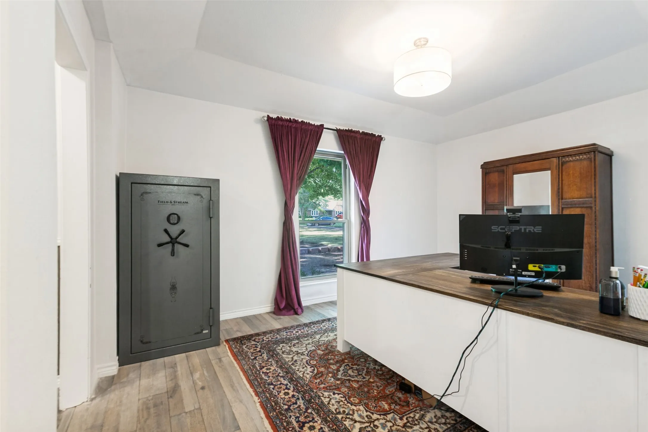 Tray ceiling, wood-finish flooring, built-in desk with wood-finish top, drum pendant light fixture, window with exterior landscaping