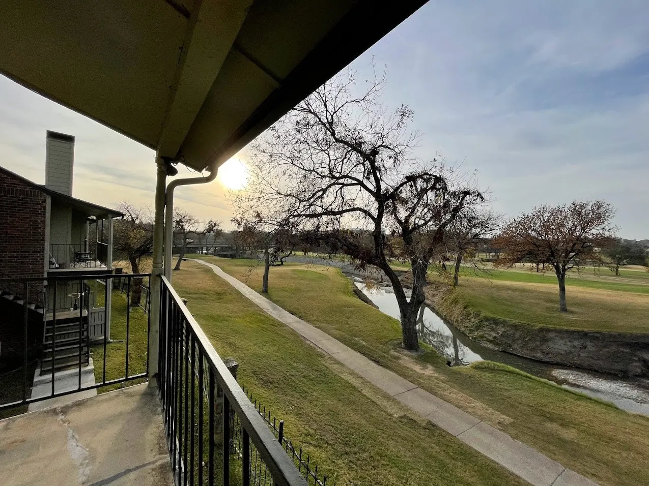 Exterior balcony with black metal railing overlooking a landscaped green space with a paved pathway and a natural creek