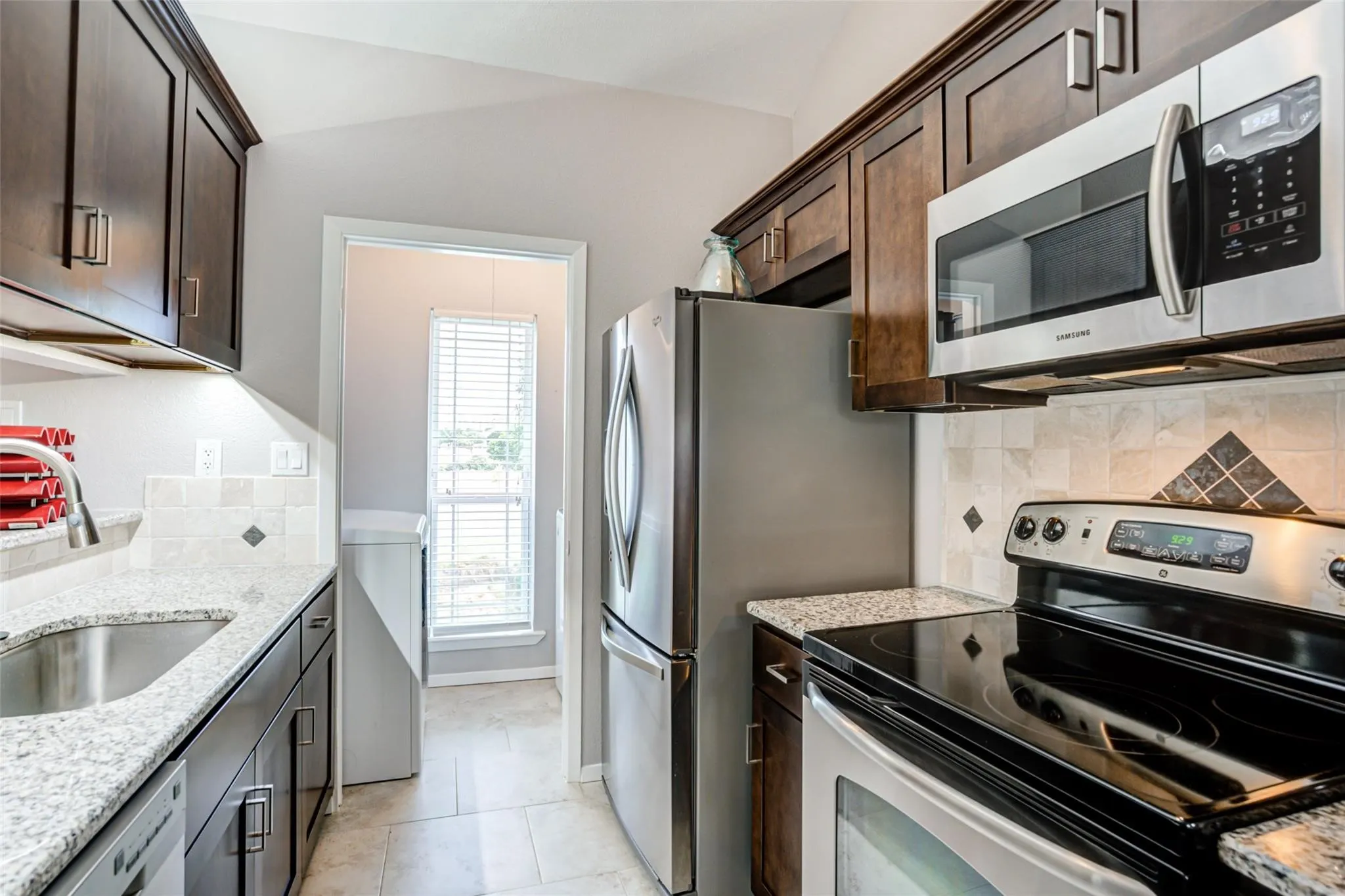 Kitchen featuring dark wood cabinetry, granite countertops, stainless steel appliances, a tile backsplash, and a vaulted ceiling