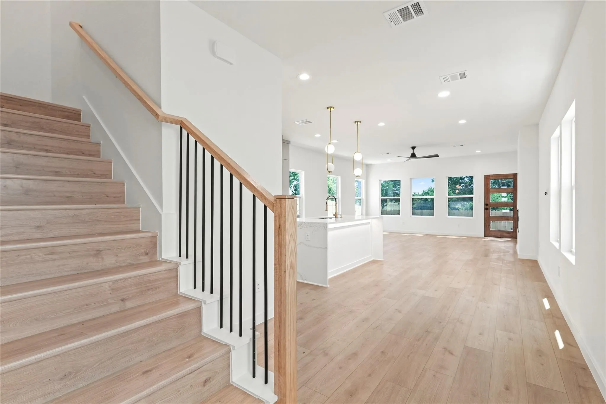 Light wood-finish flooring, staircase with wood handrail and black balusters, recessed lighting, white kitchen island with undermount sink and pendant lighting, and a dark wood entry door