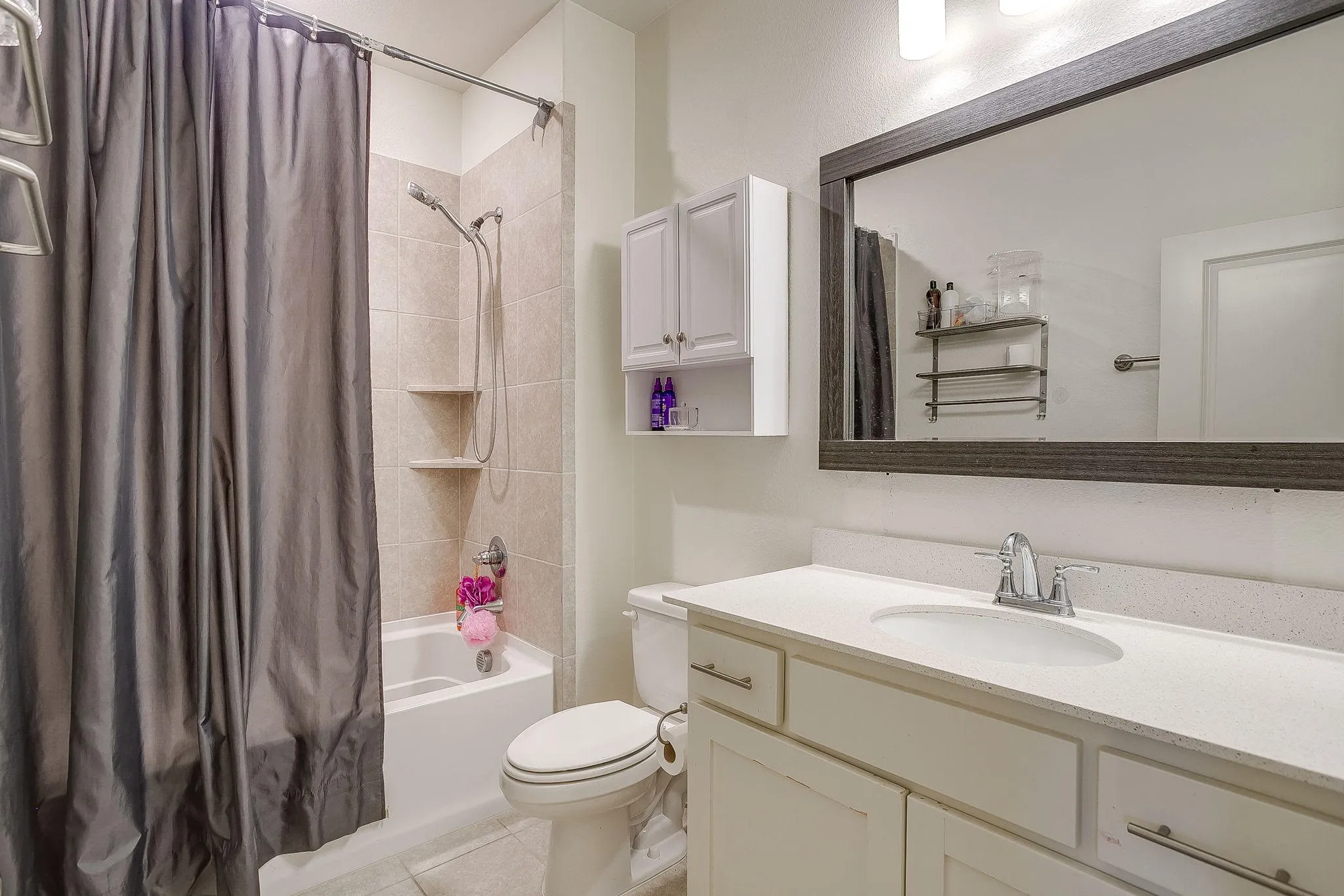 Bathroom featuring a shower-tub combination with tiled surround and built-in shelving, a single vanity with a white countertop, and an overhead mirror with a dark frame