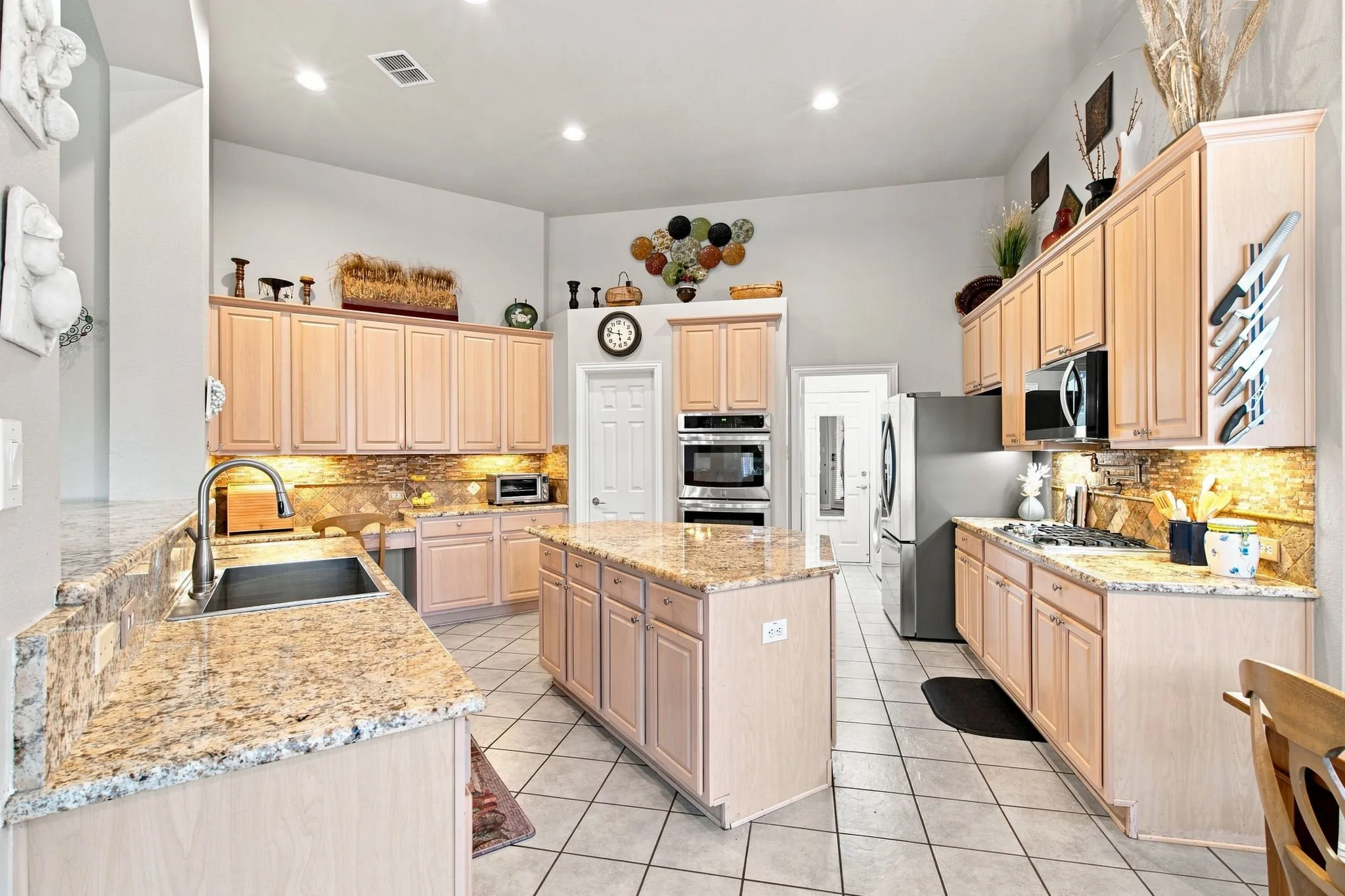 Kitchen featuring an island, light wood-finish cabinetry, built-in stainless steel appliances, granite-style countertops, and tiled flooring