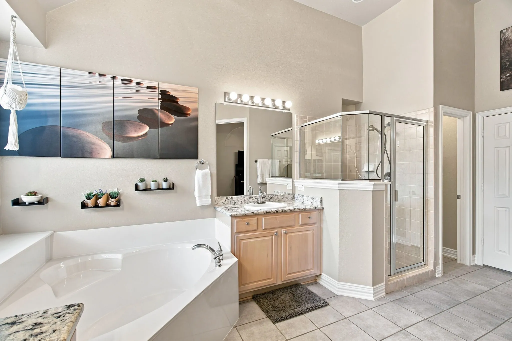 Corner soaking tub, separate glass-enclosed shower, light wood-finish vanity, light-toned stone-finish countertop, and tile flooring