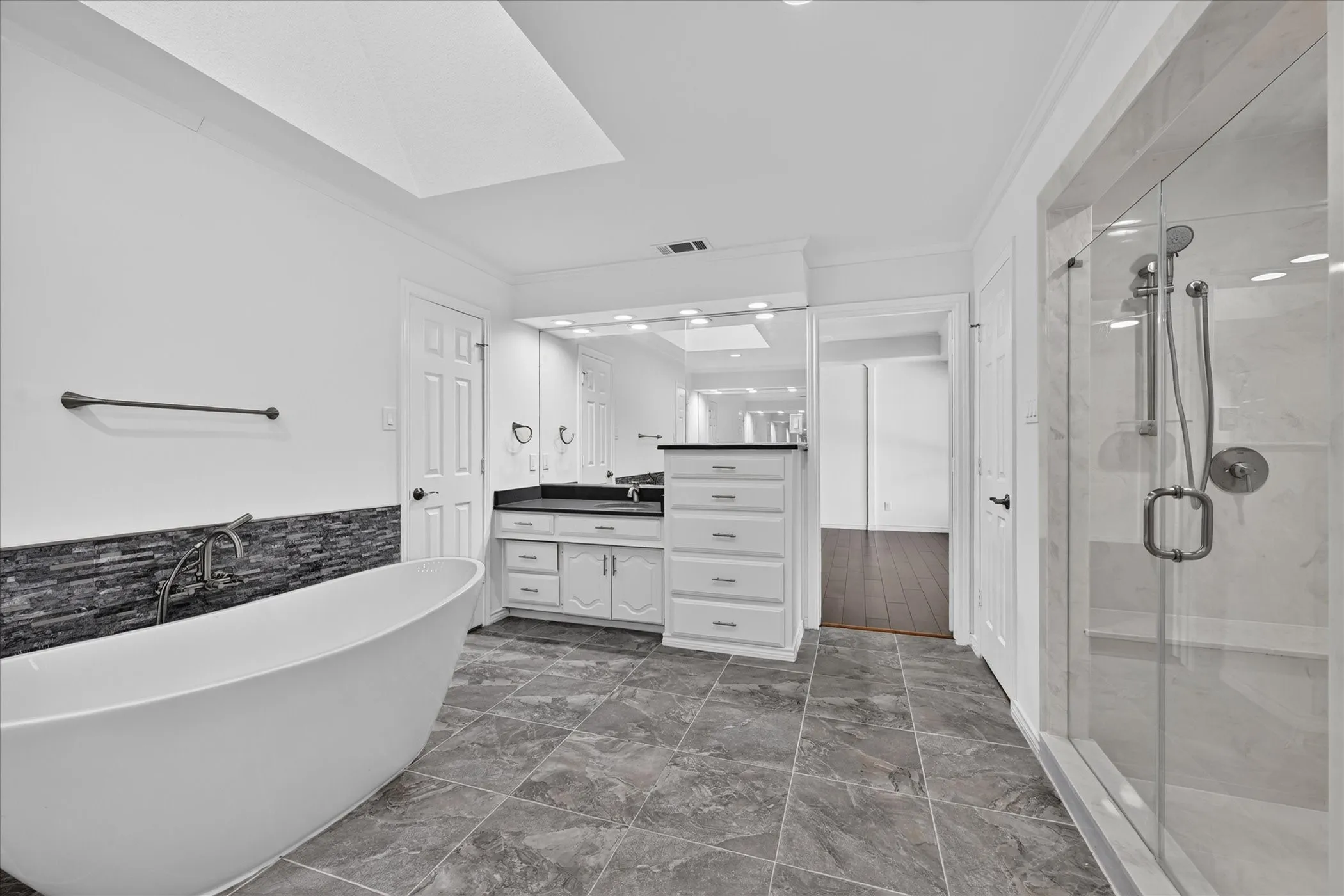 Freestanding soaking tub with a stacked stone tile accent wall, glass-enclosed shower, dual vanity with undermount sinks, and stone countertop