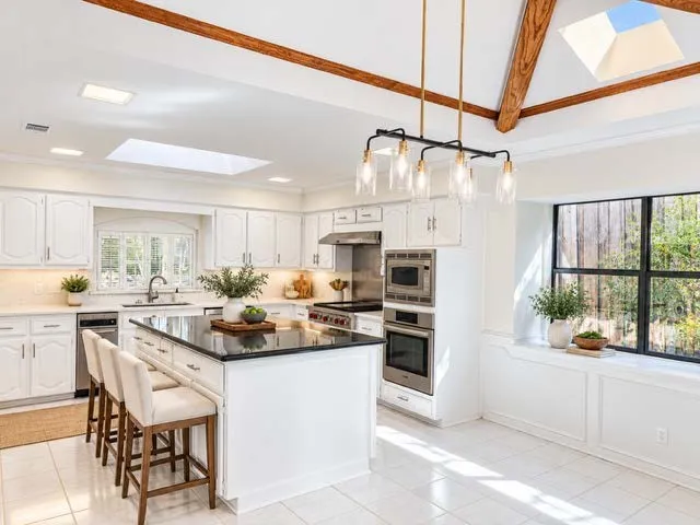staged spacious kitchen with exposed wood beams, multiple skylights, a center island with a dark countertop, stainless steel appliances, and white cabinetry