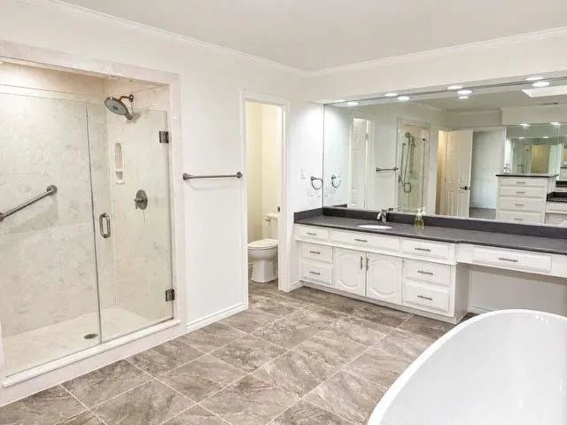 Glass-enclosed shower with stone-finish tile, extensive vanity with dual sinks, built-in makeup counter, and large wall-mounted mirror