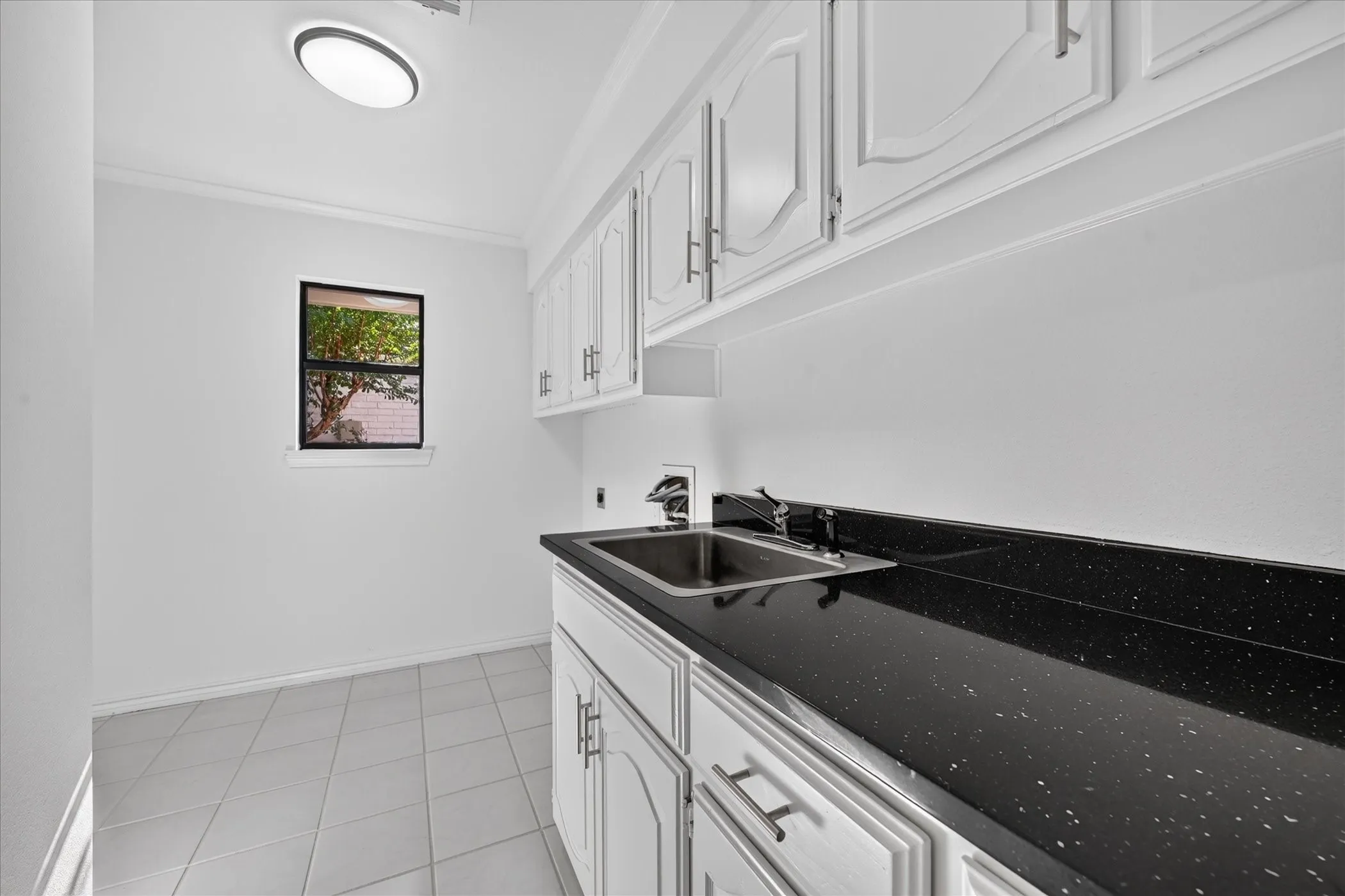 Built-in cabinetry with white paneling and silver-tone hardware, black speckled countertop, stainless steel sink, tiled flooring, and a window providing natural light, much cabinet space, extra room for freezer