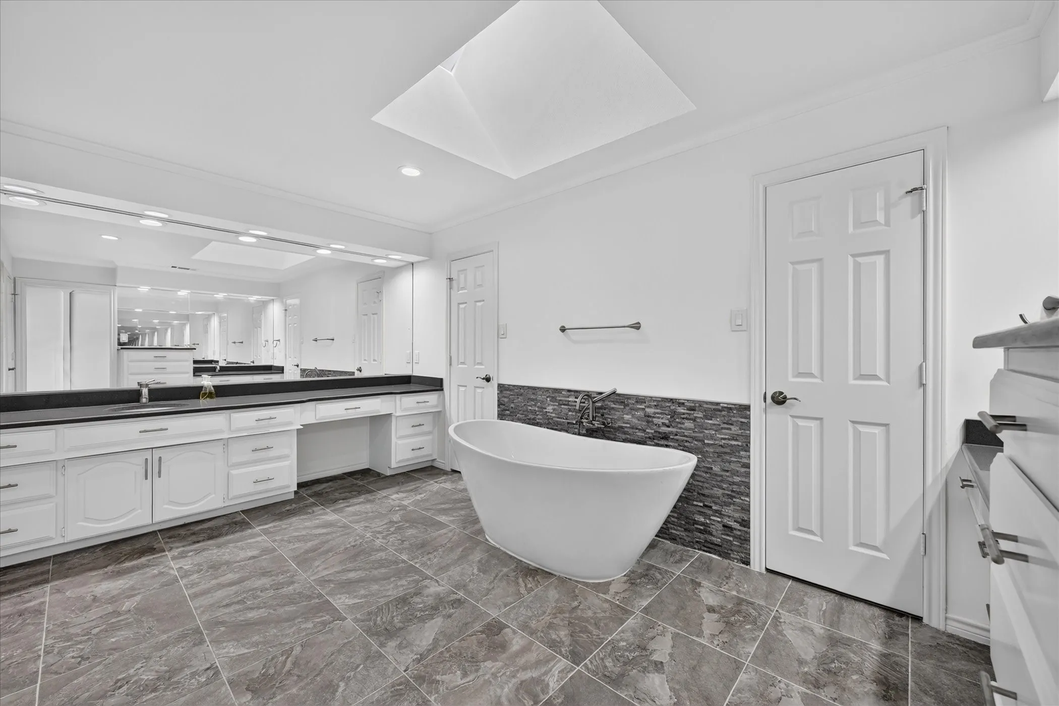 Freestanding soaking tub, skylight, extensive vanity with dual sinks, dark stone countertops, and large format floor tiling