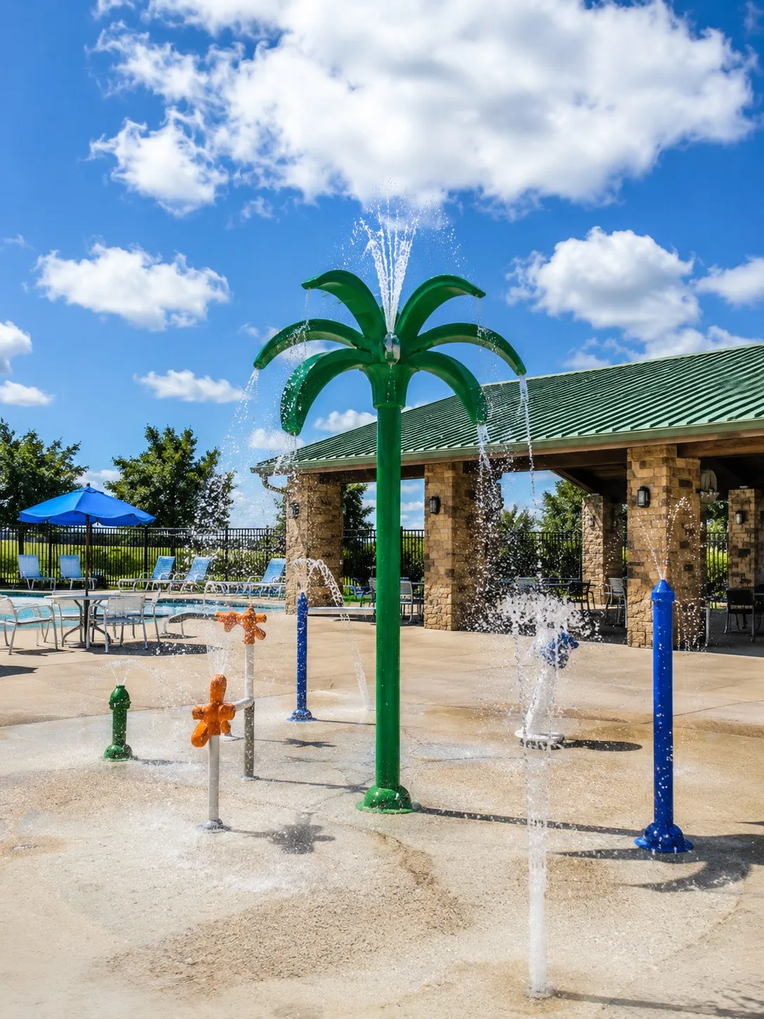 Splash pad with multiple water features, adjacent covered patio with stone columns, and nearby swimming pool with lounge seating
