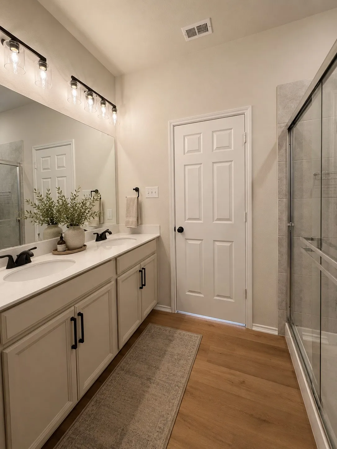 Dual sink vanity with white countertop, black hardware, wood-finish flooring, and a glass-enclosed shower