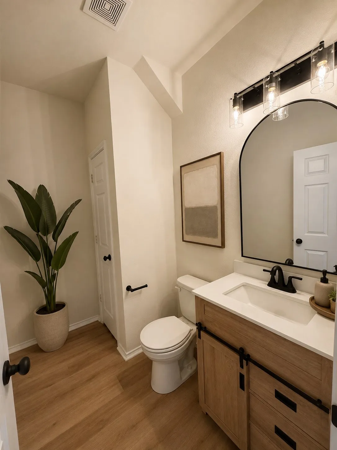 Light wood-finish flooring, single vanity with light countertop, black fixtures, and arched mirror