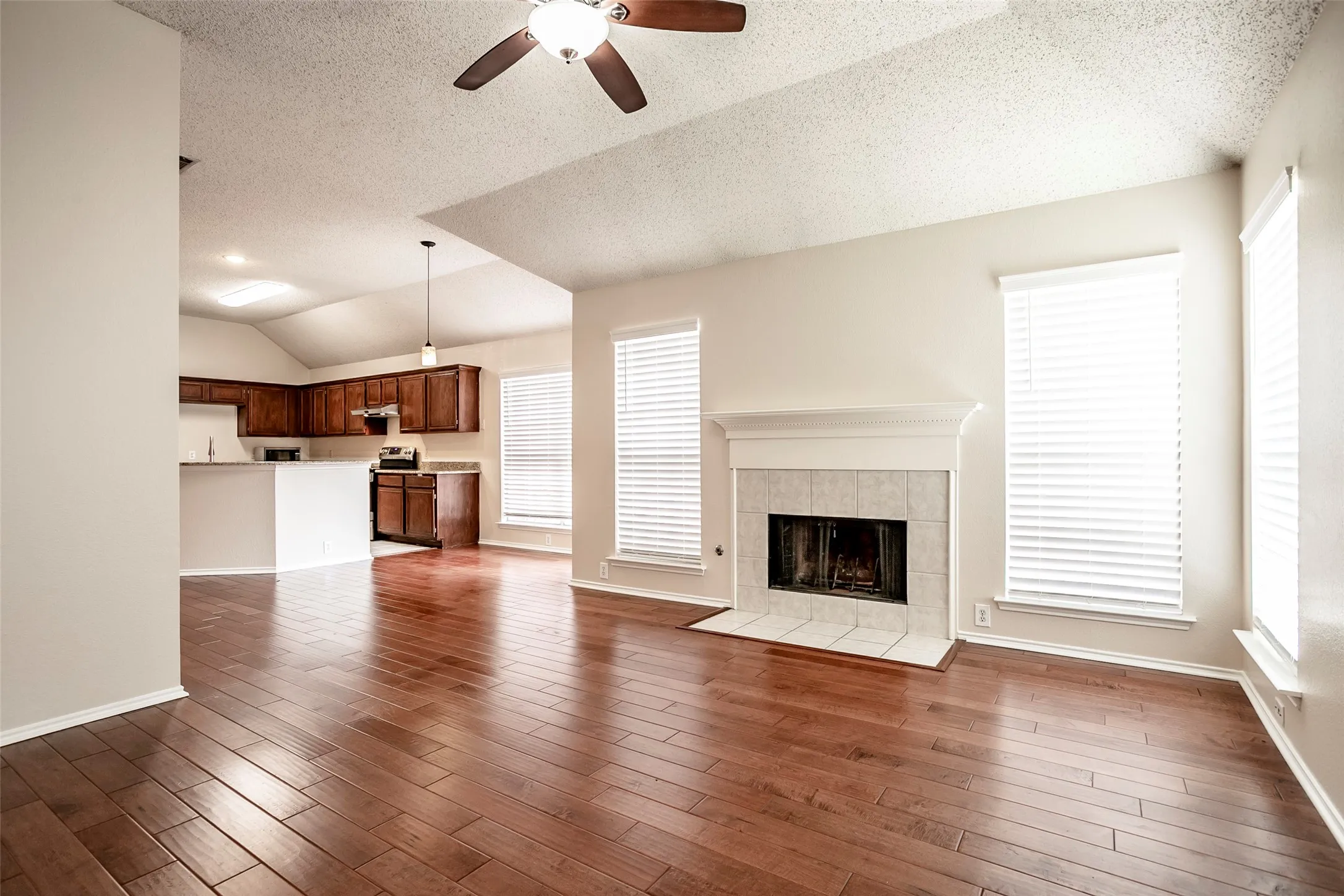 Open-concept layout with wood-finish flooring, a tiled fireplace with mantel, and a ceiling fan