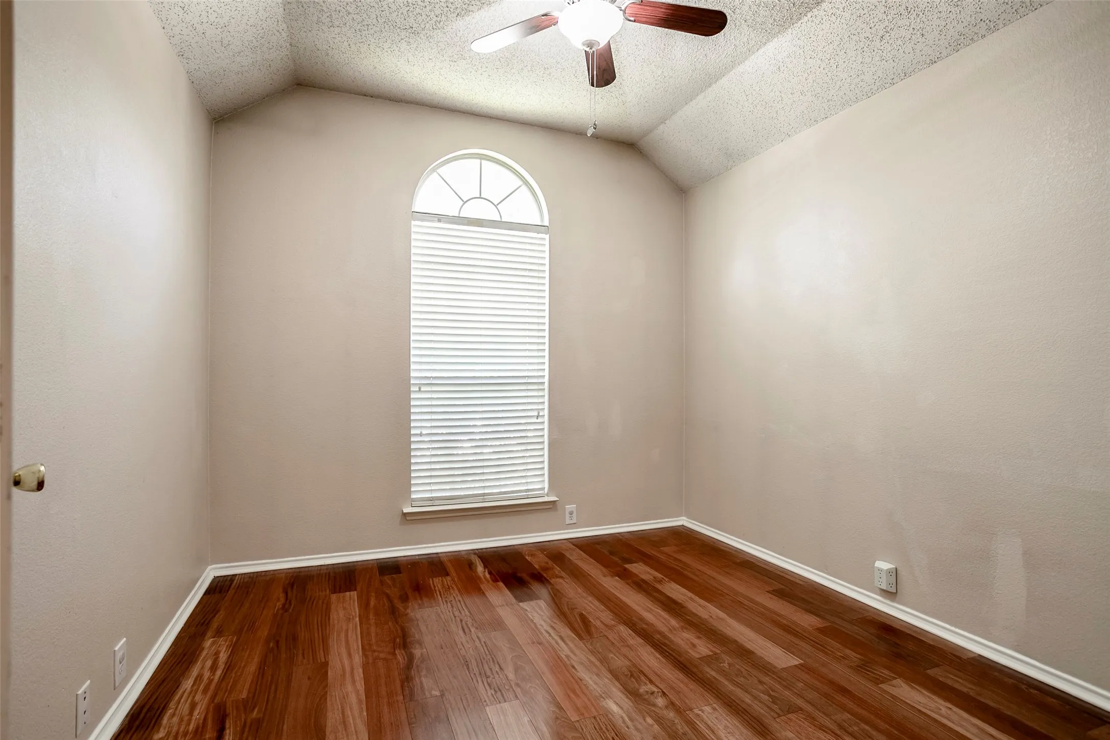 Arched window with blinds, vaulted ceiling, ceiling fan with light fixture, light-toned walls, and wood-finish flooring