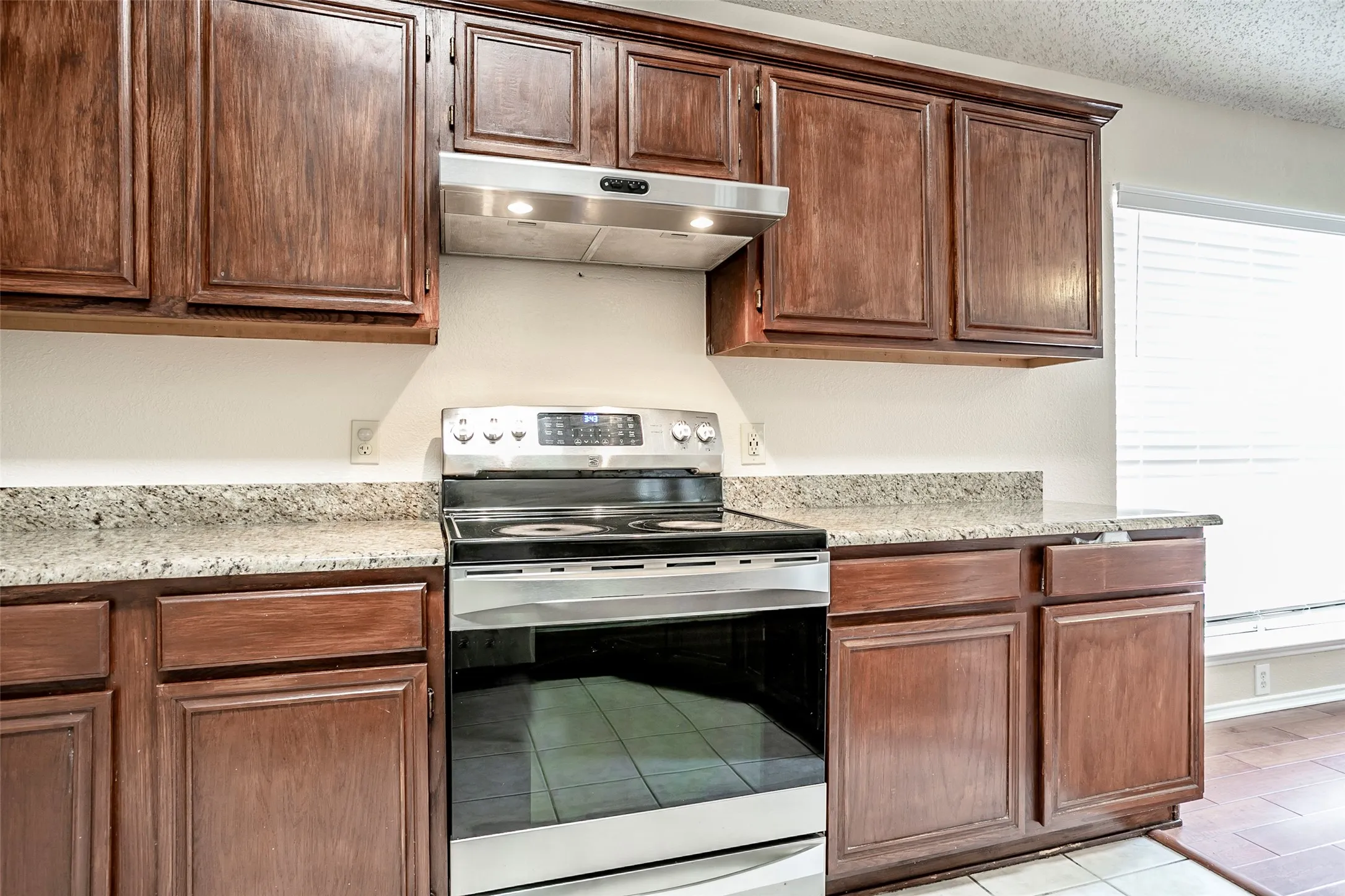 Stainless steel range, stainless steel range hood, wood-finish cabinetry, speckled countertops, and a window with blinds