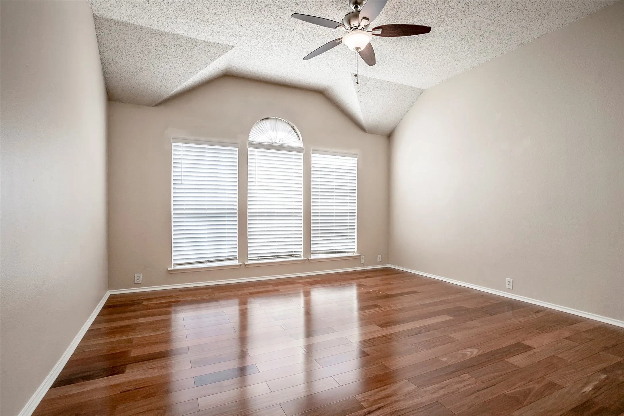 Vaulted ceiling, ceiling fan with integrated lighting, wood-finish flooring, three large windows, and a palladian window