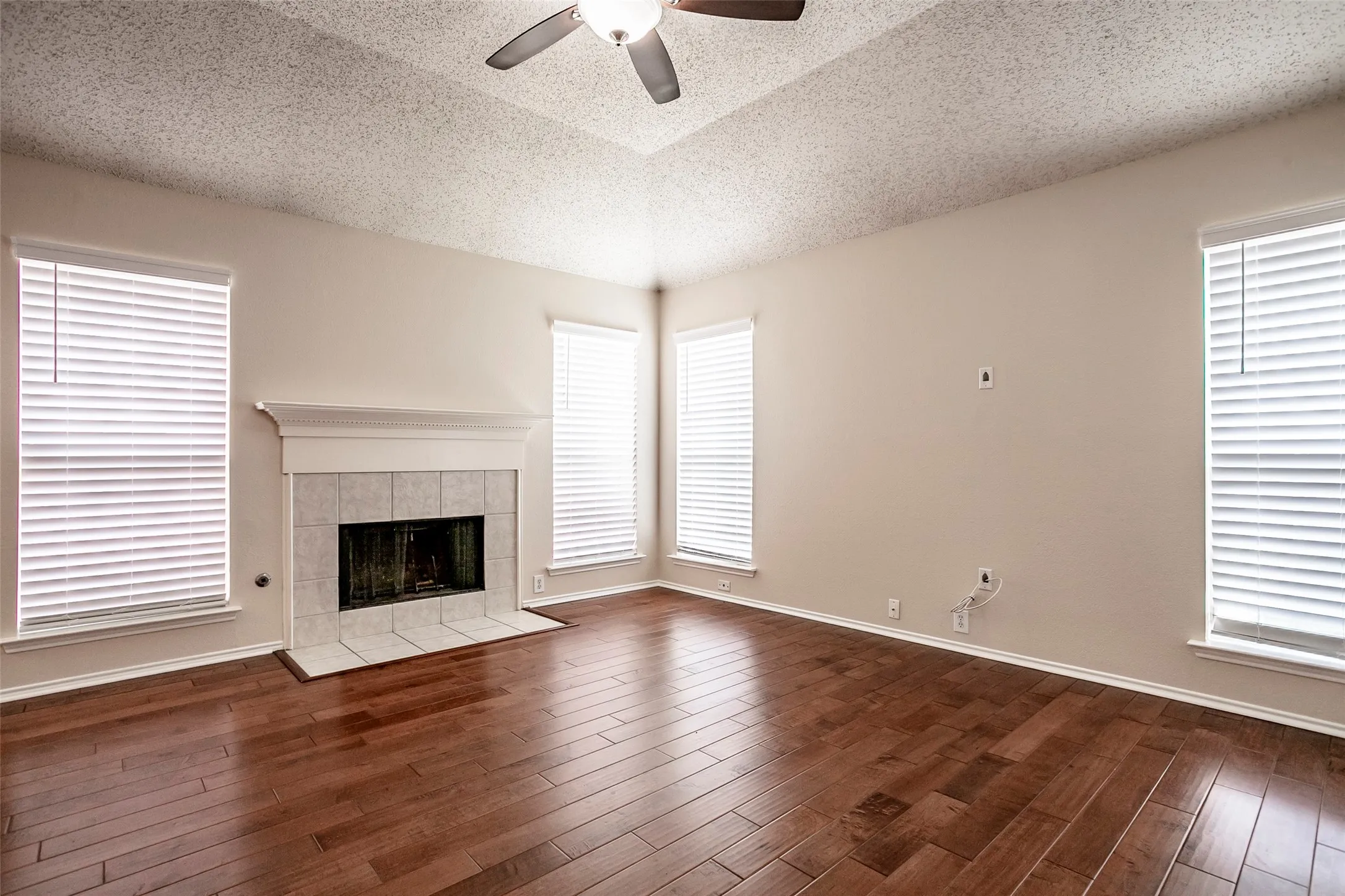 Wood-finish flooring, tiled fireplace with white mantel, ceiling fan, and multiple windows with blinds