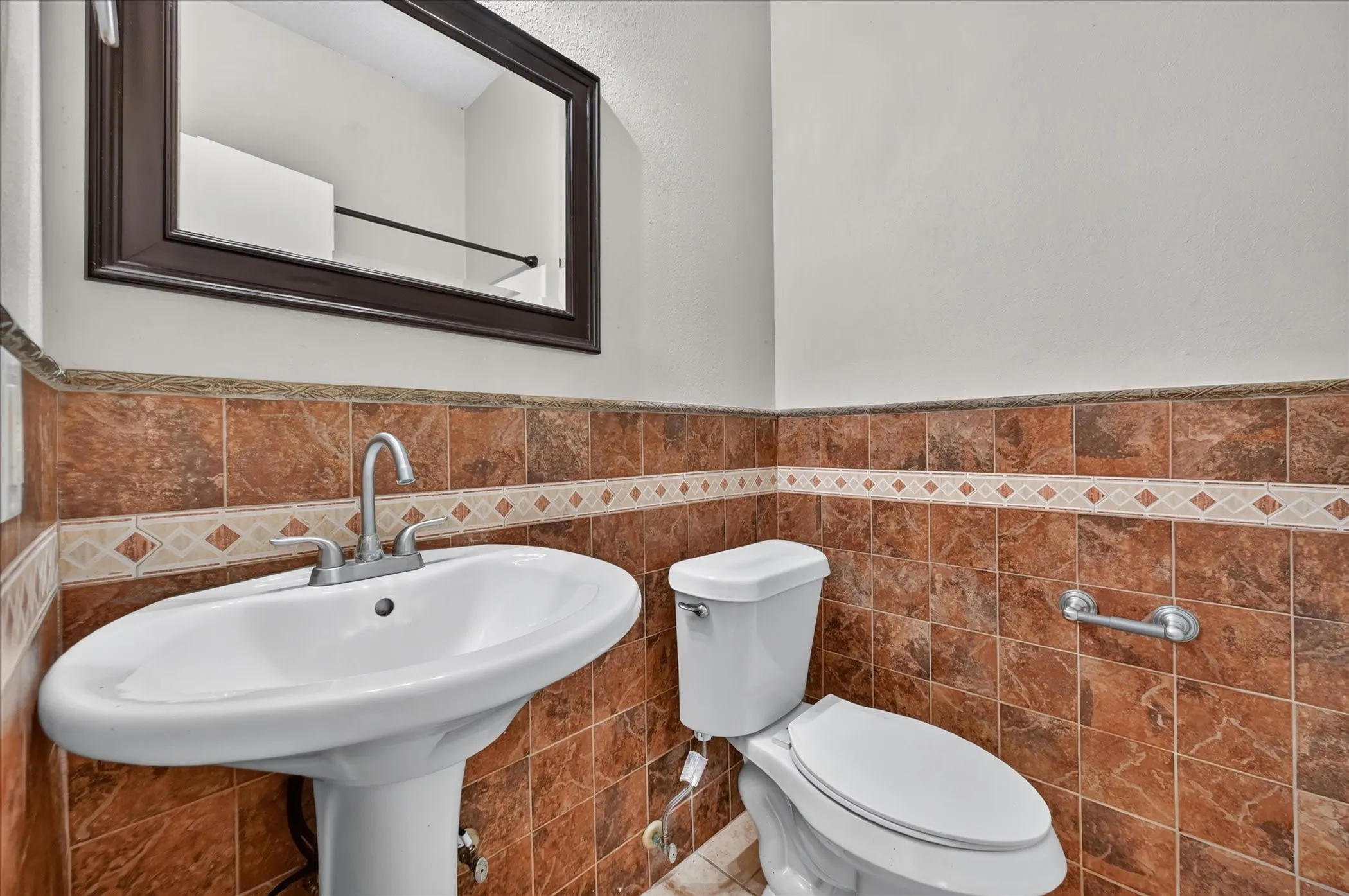 Pedestal sink with chrome faucet, framed mirror, and tiled walls featuring a decorative border