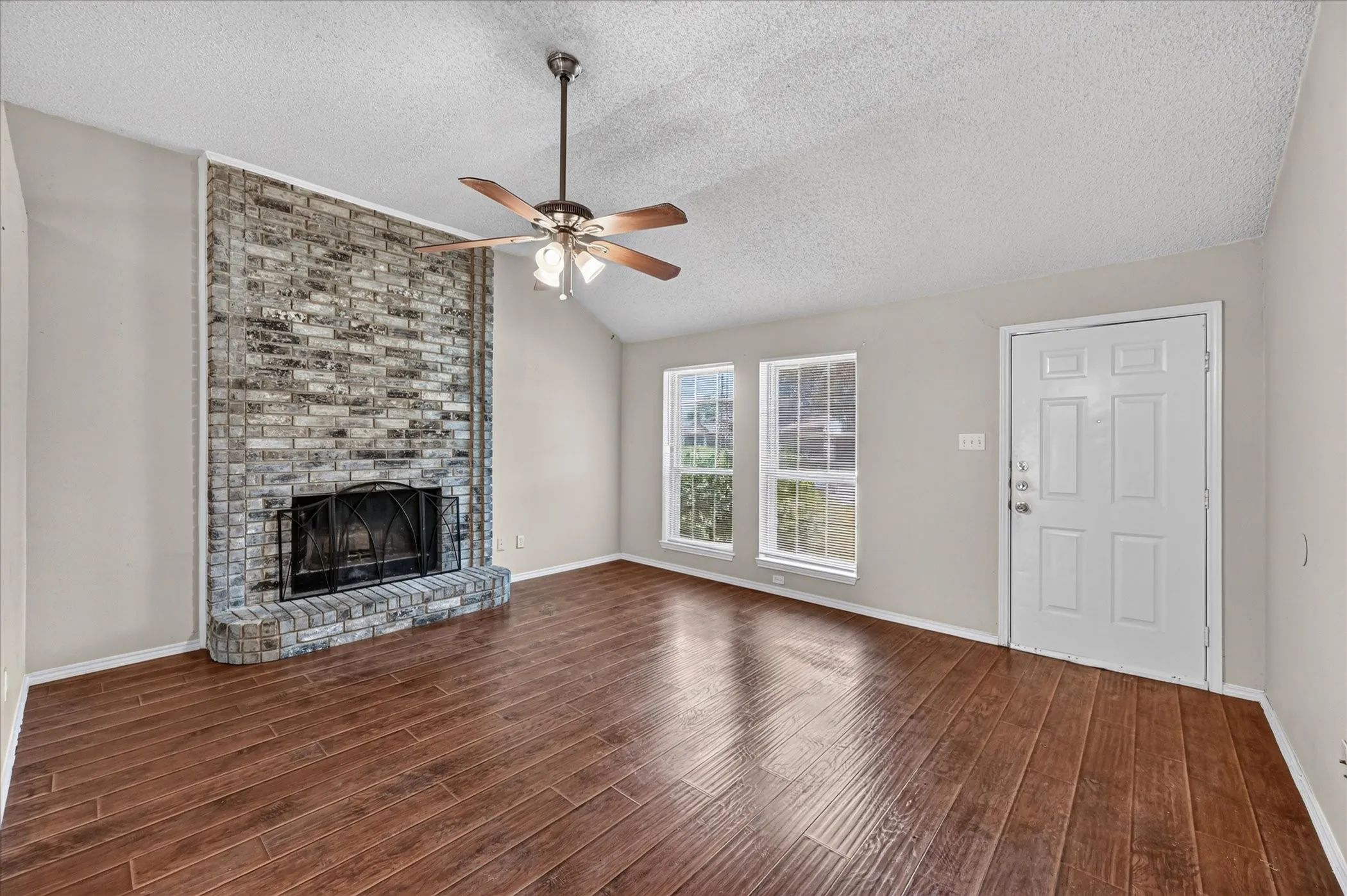 Vaulted ceiling with a ceiling fan, brick fireplace, wood-finish flooring, and multiple windows