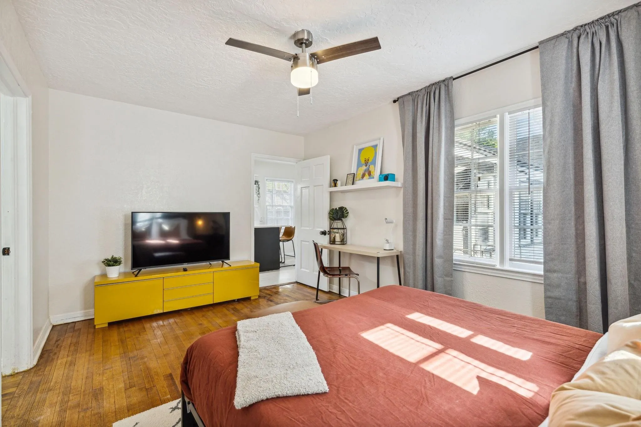 Wood-finish flooring, ceiling fan with integrated lighting, window with blinds, and white floating shelf