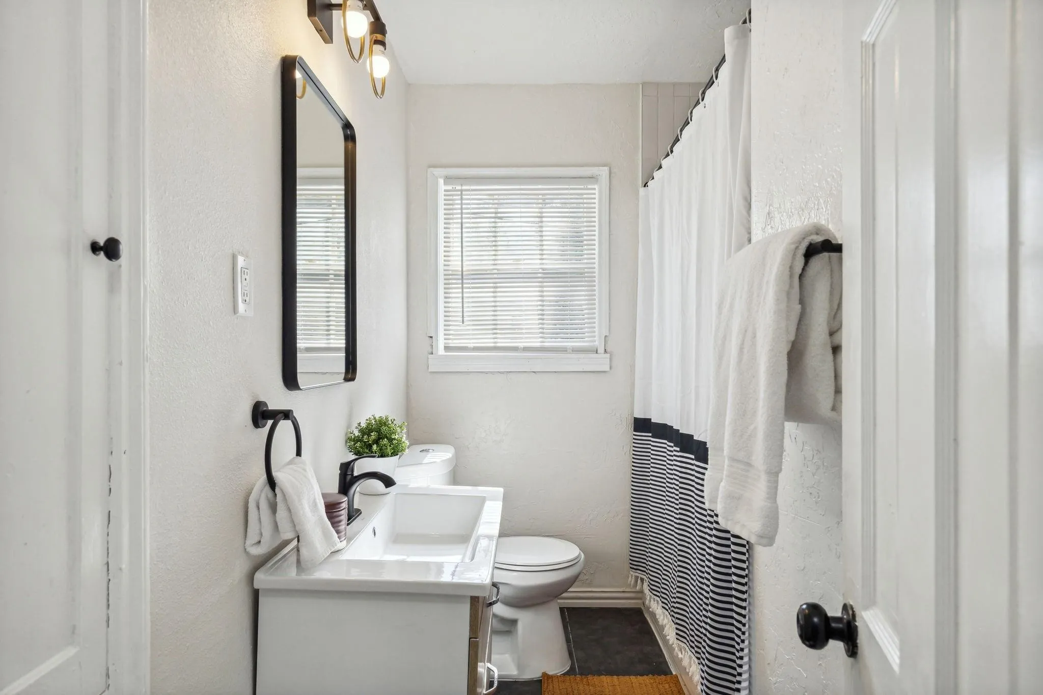 Bathroom featuring a single vanity, wall-mounted mirror, overhead lighting, window with blinds, and shower with a striped curtain